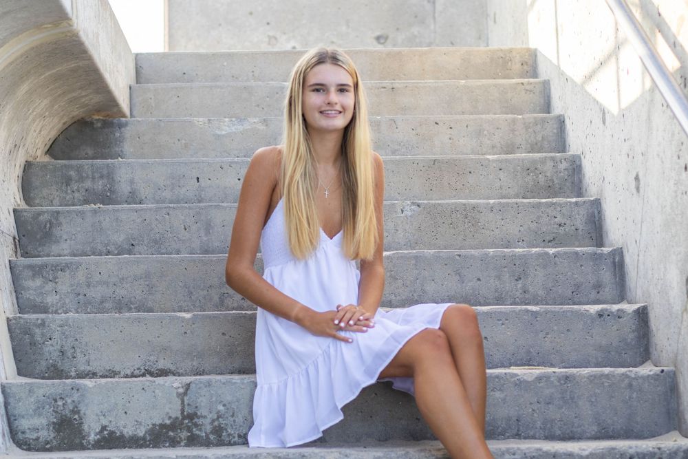 Smiling young blonde woman in white dress sitting on concrete steps outdoors.