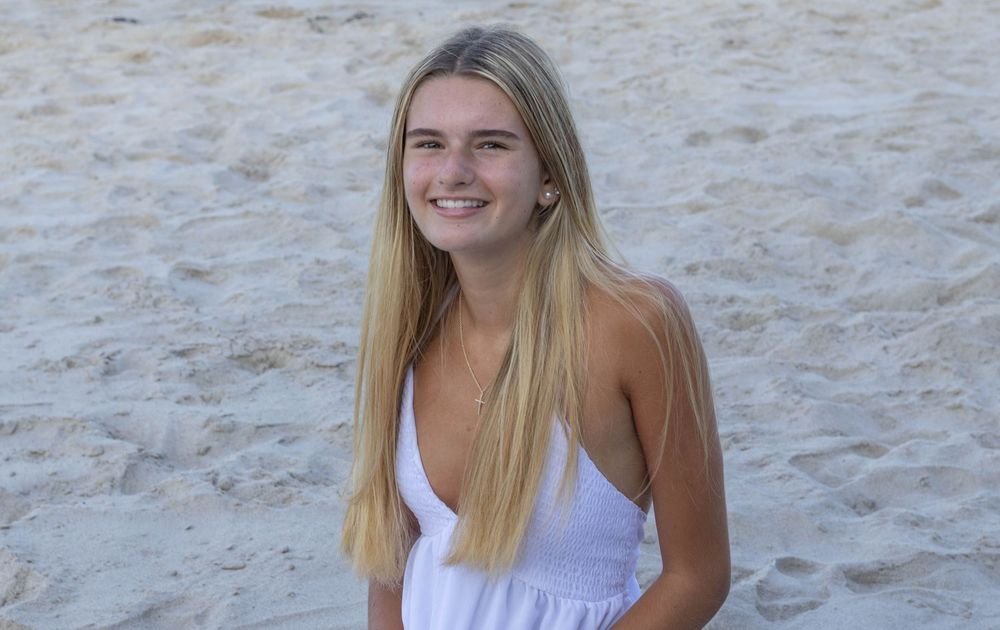 Smiling young blonde woman in white dress sitting on sandy beach.