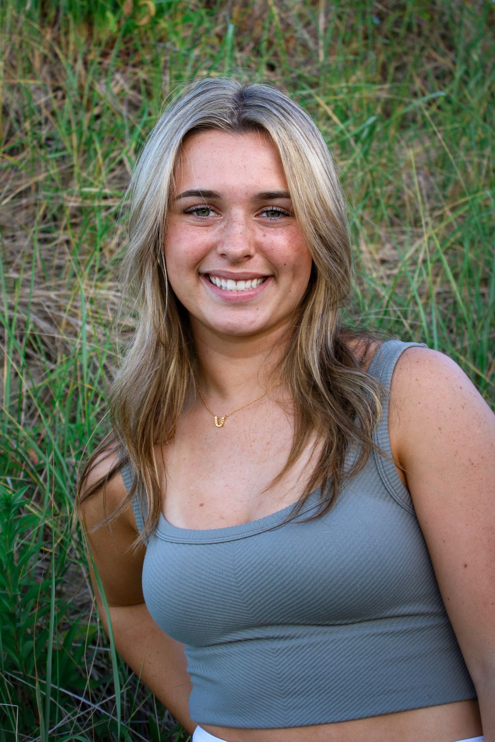 Smiling young blonde woman in gray crop top sitting in a green grassy field.