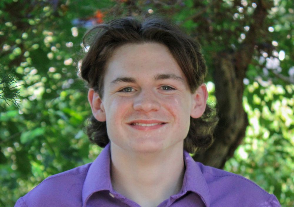 Smiling teenage boy in a purple polo shirt posing outdoors with green foliage background.