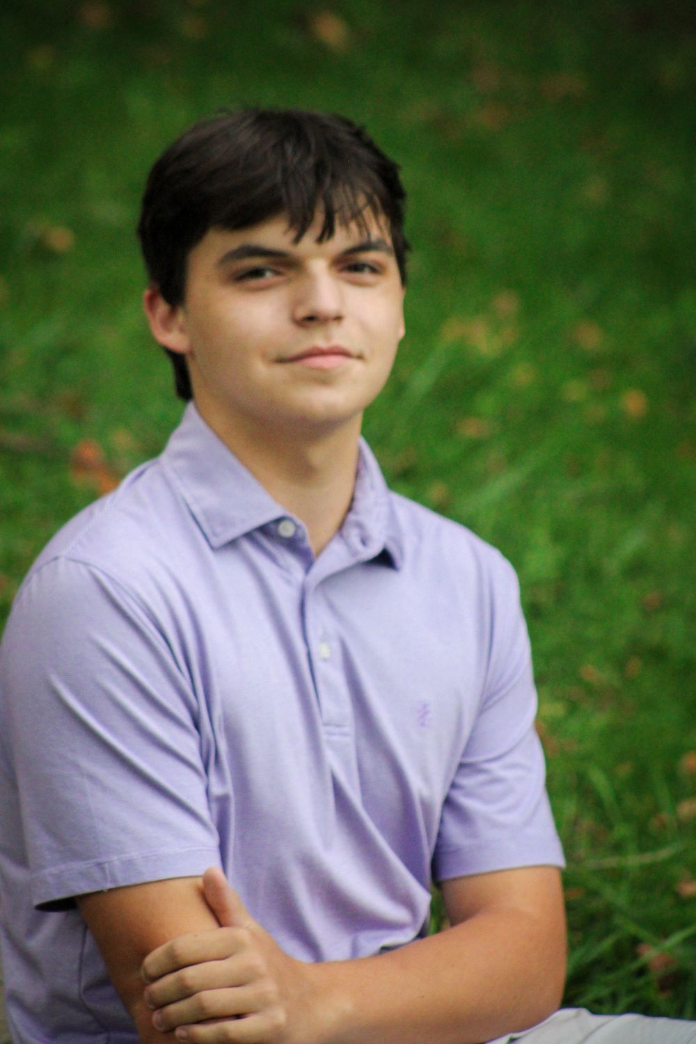 Close-up portrait of a teenage boy in a lavender polo shirt sitting on green grass outdoors.