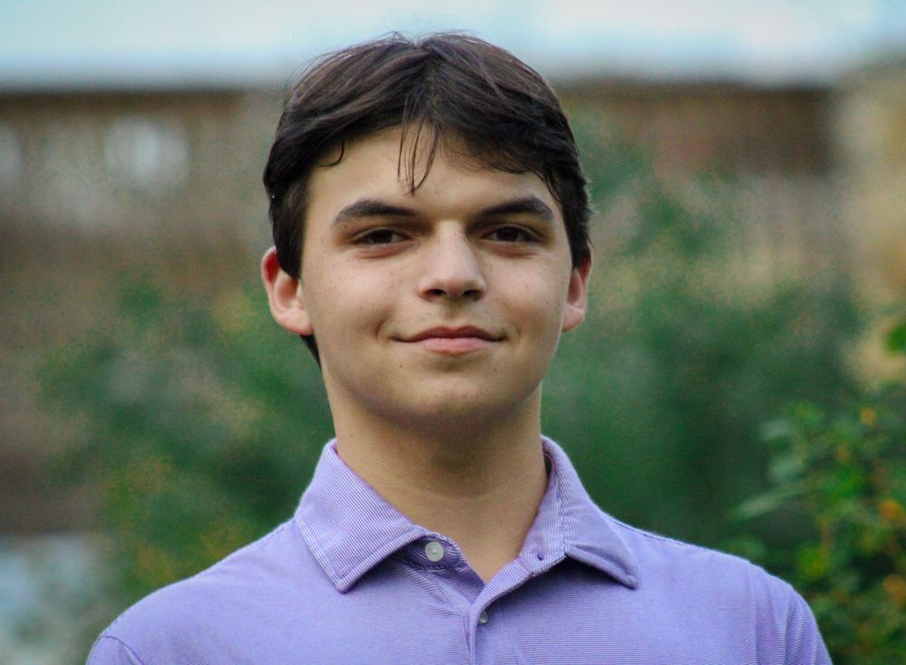 Close-up portrait of a smiling teenage boy wearing a lavender polo shirt outdoors.
