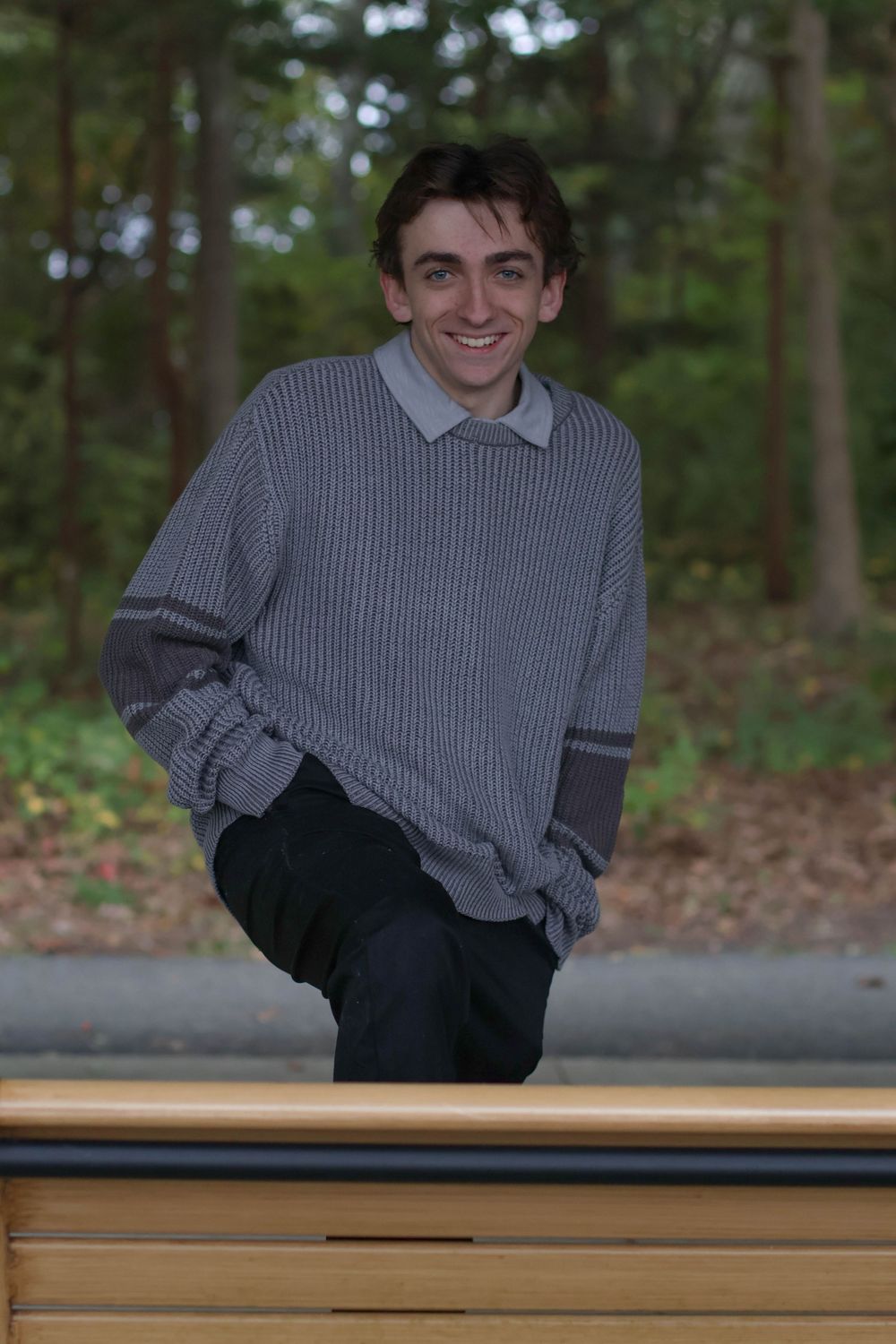 Smiling young man in gray striped sweater leaning on bench in wooded park.