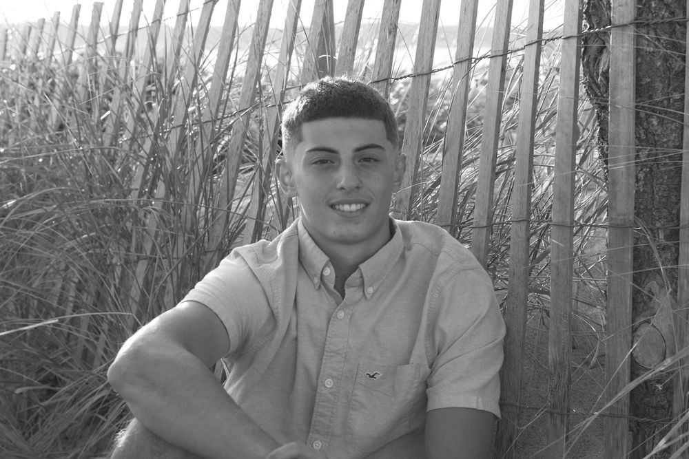 Young man smiling in black and white photo near wooden fence and beach grass.