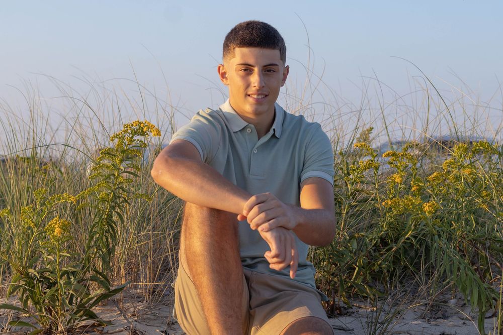 Young man in sage polo sitting on sand among wildflowers at the beach.