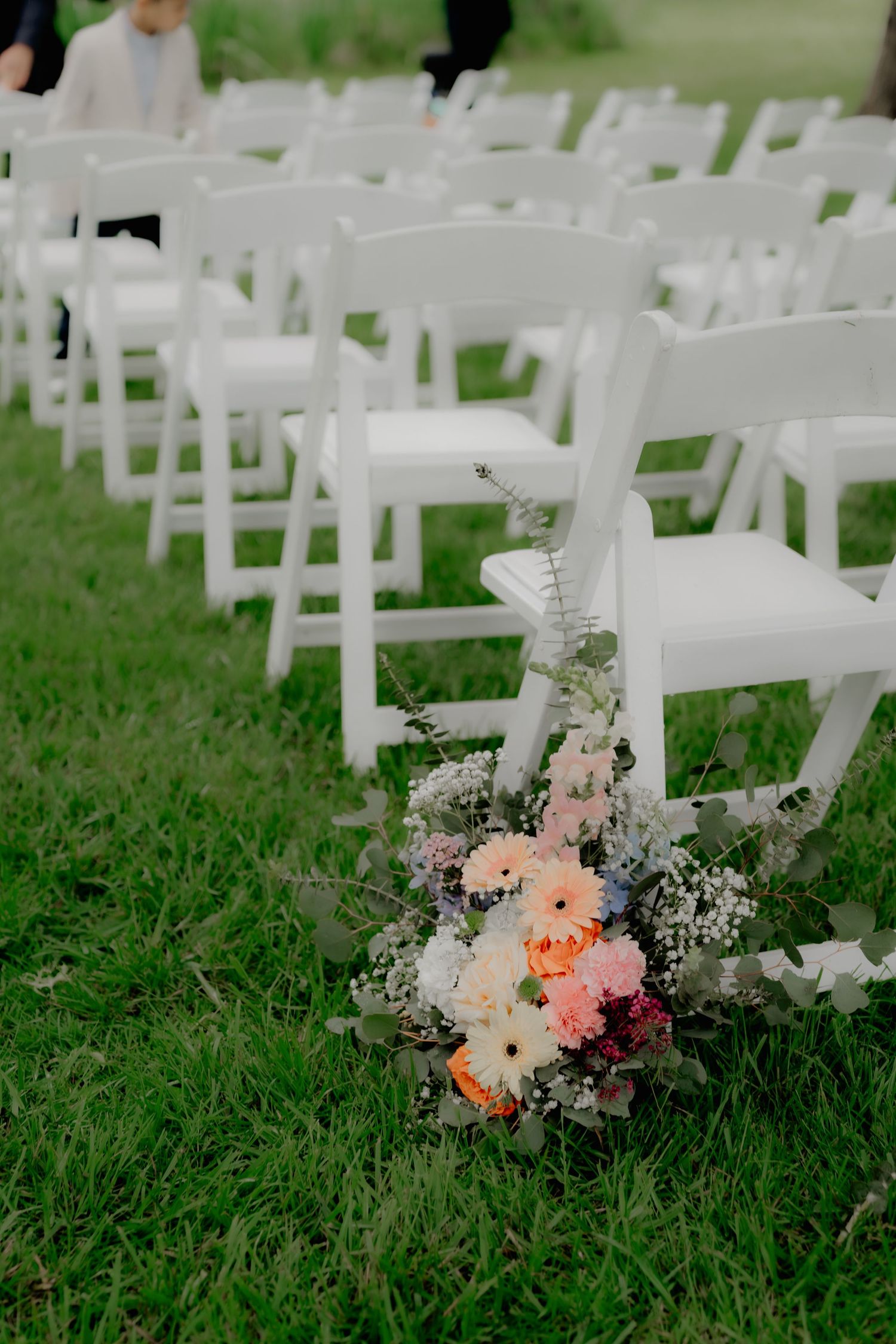 White folding chairs arranged on green grass with a colorful floral bouquet at the aisle for an outdoor wedding.