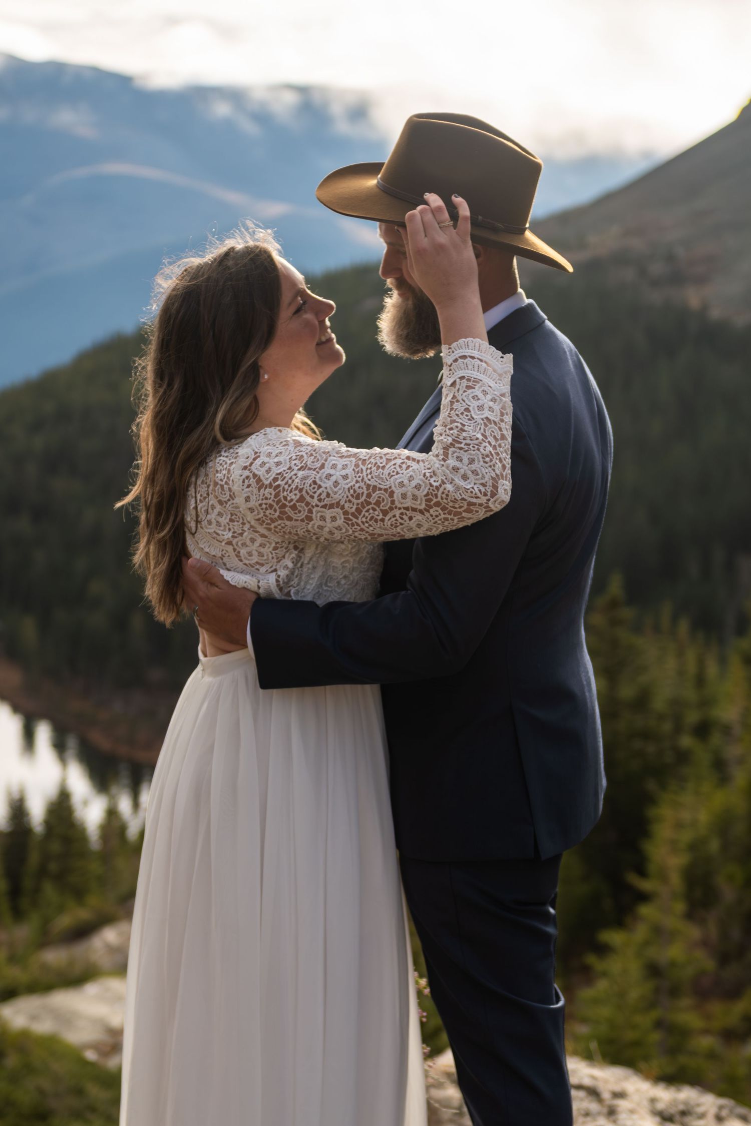 A bride and groom embrace on a mountain overlook, sharing a romantic moment in wedding attire with scenic forest views.