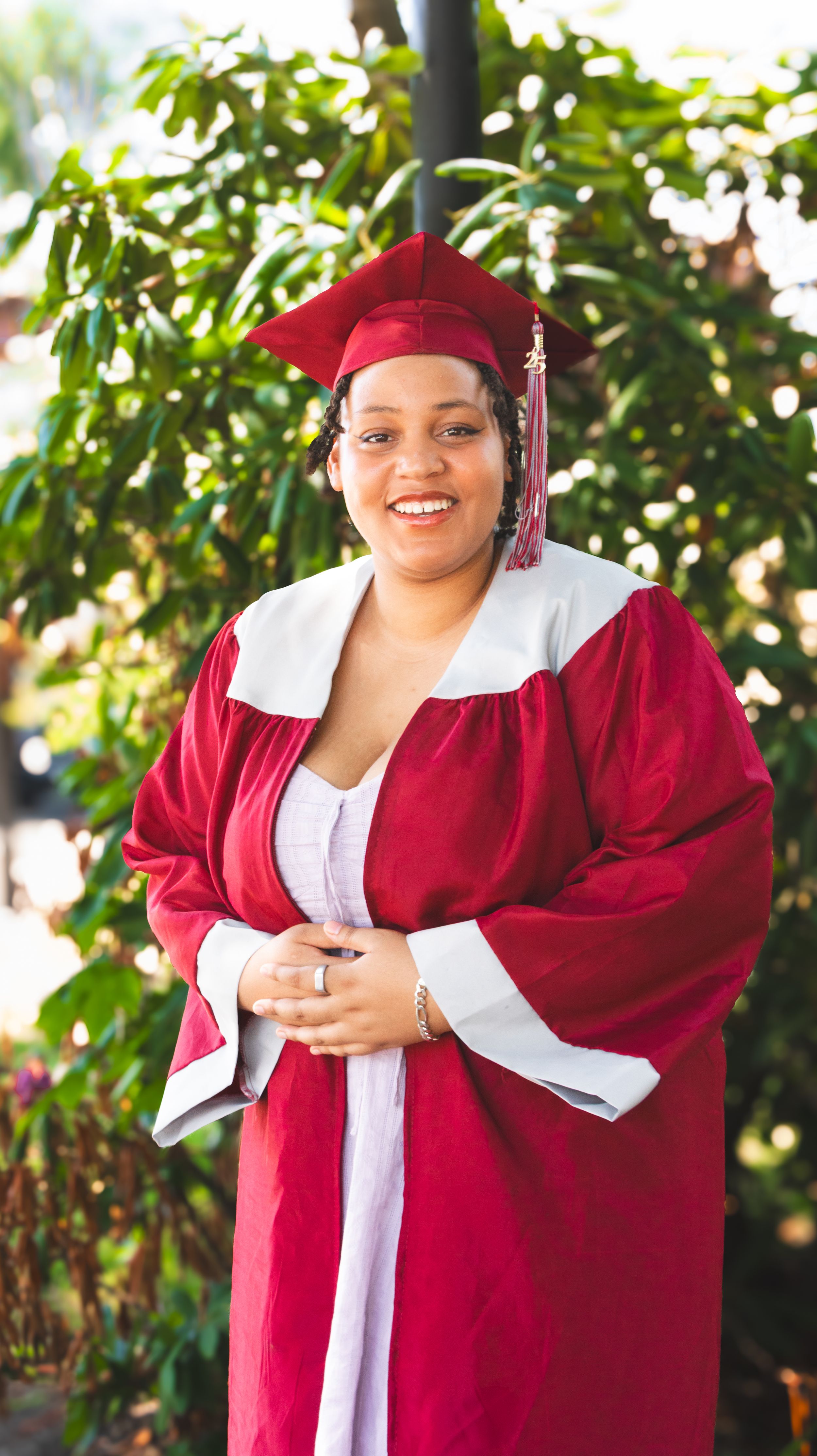 Smiling graduate in red cap and gown poses outdoors in front of green trees on graduation day.