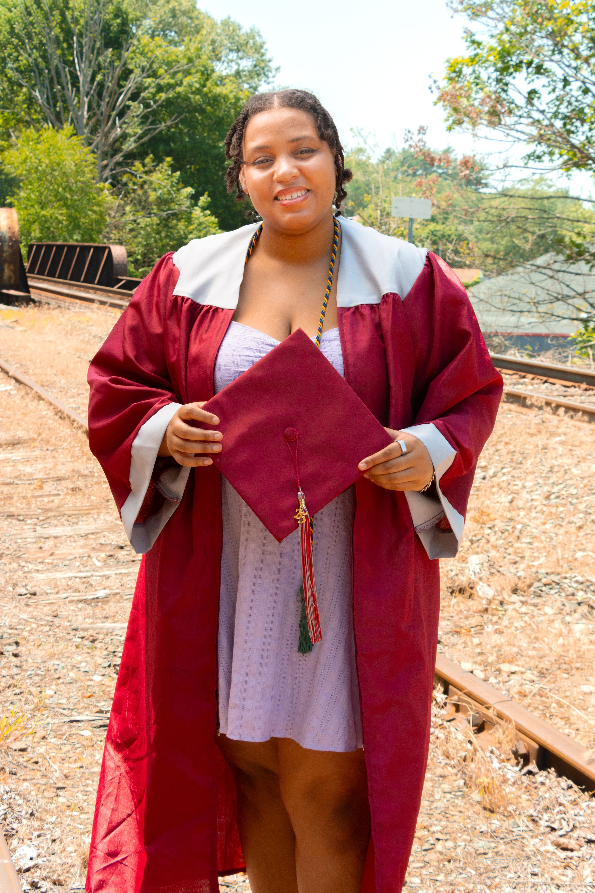 Young woman in maroon graduation gown and cap smiling outdoors near railroad tracks on a sunny day.