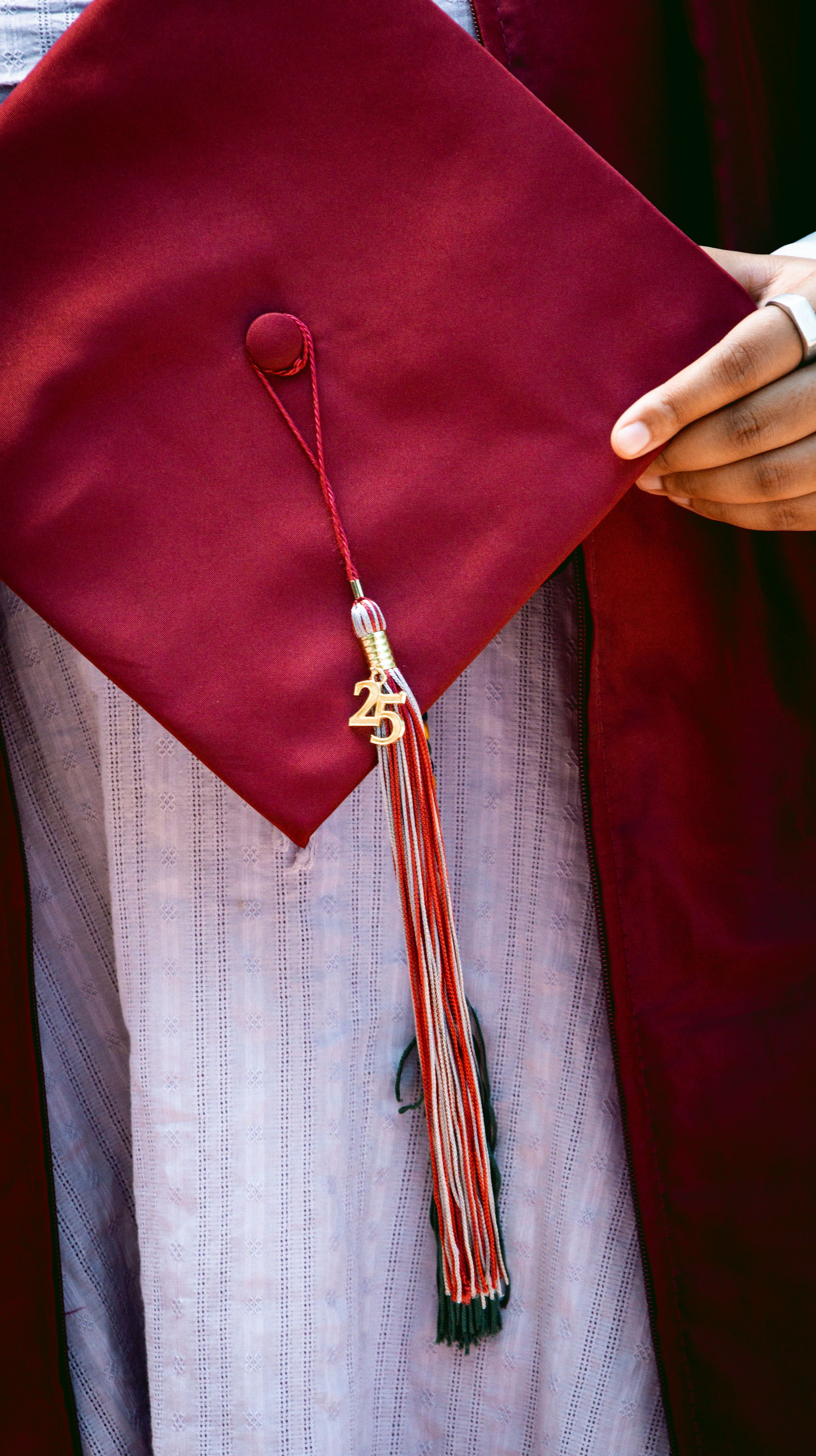 A close-up of a maroon graduation cap with a tassel featuring a gold 25 charm, held by a graduate in matching regalia.