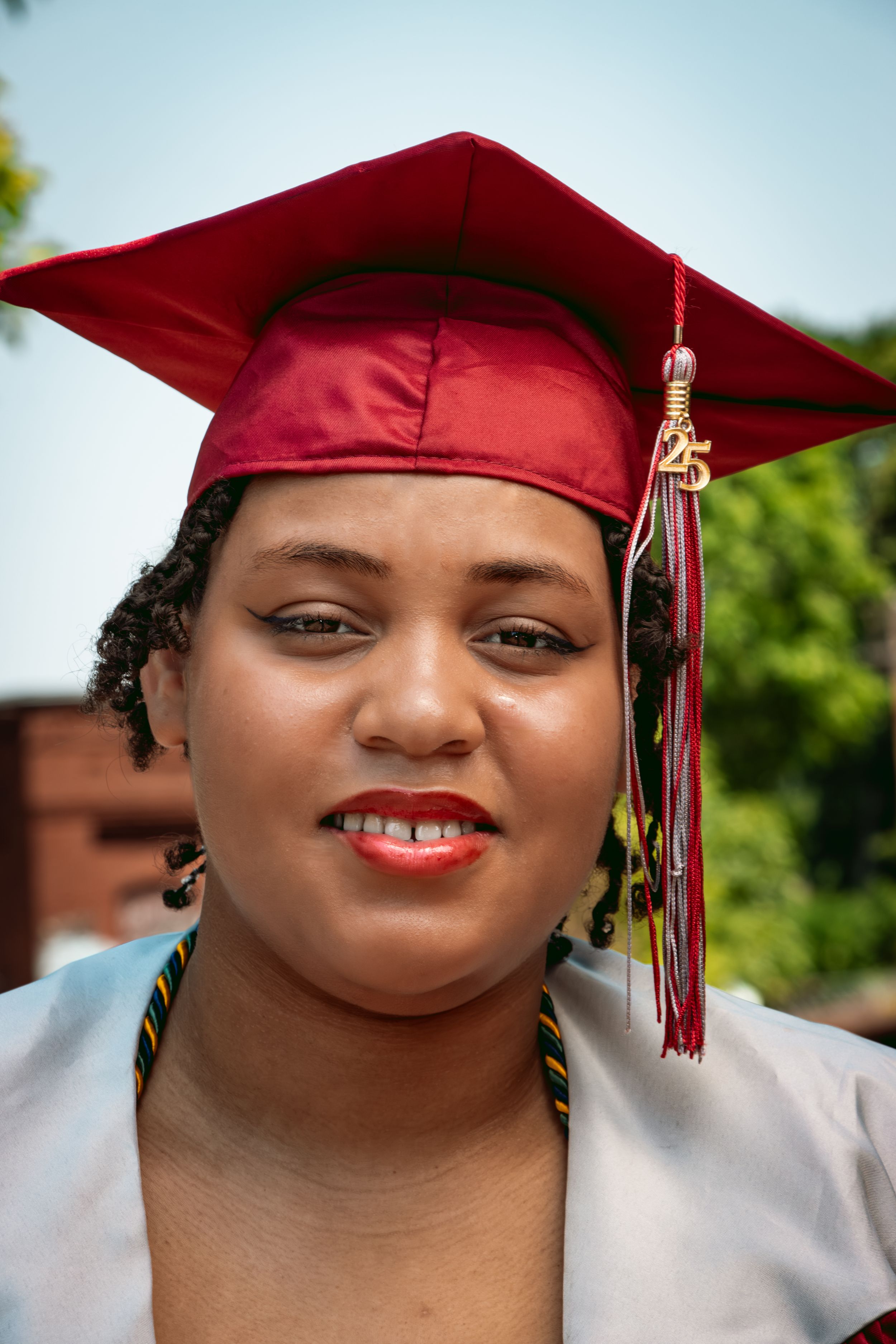Young woman in red graduation cap and gown smiling outdoors on graduation day.