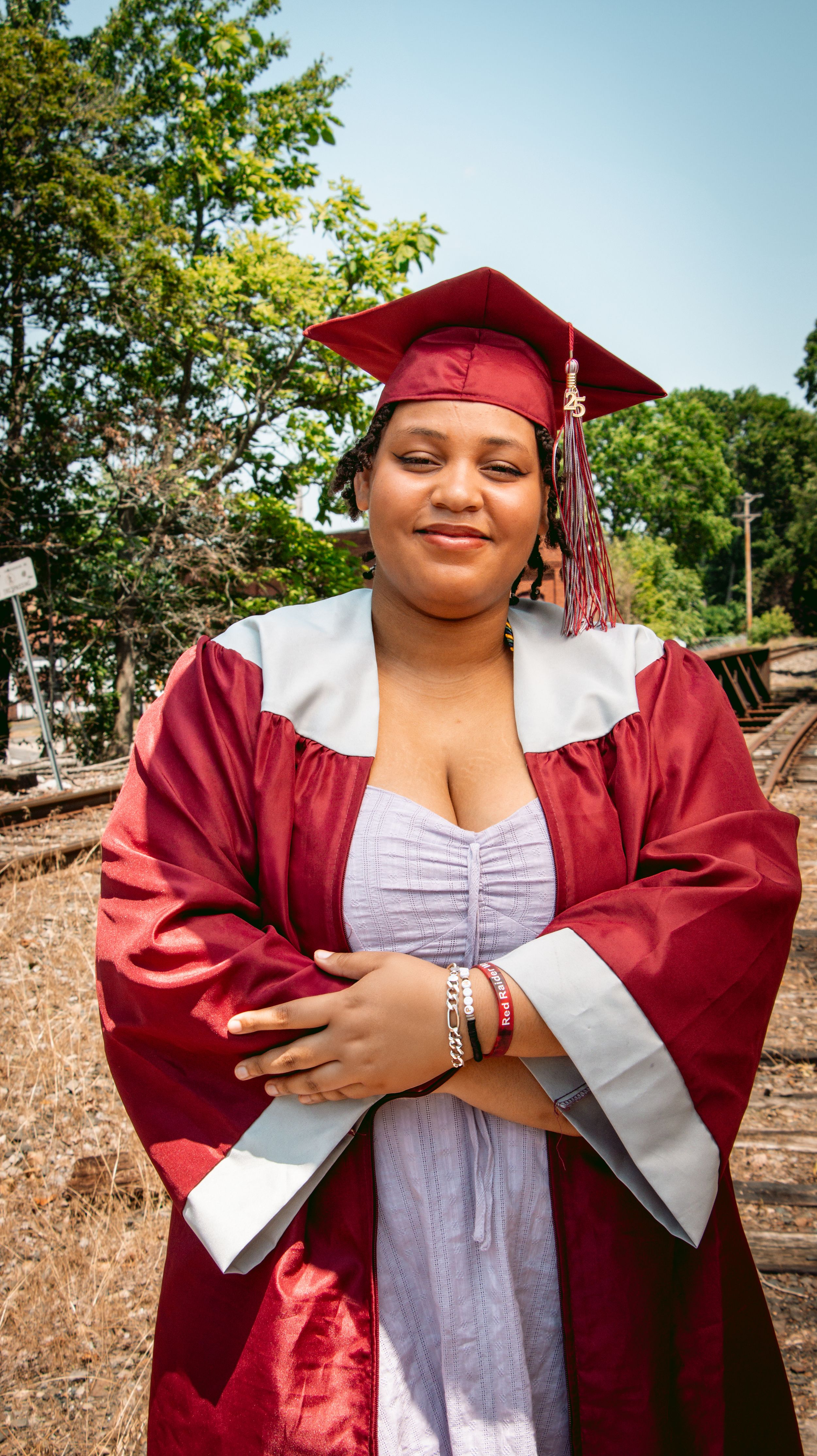 Graduate in red cap and gown posing outdoors with arms crossed on graduation day.