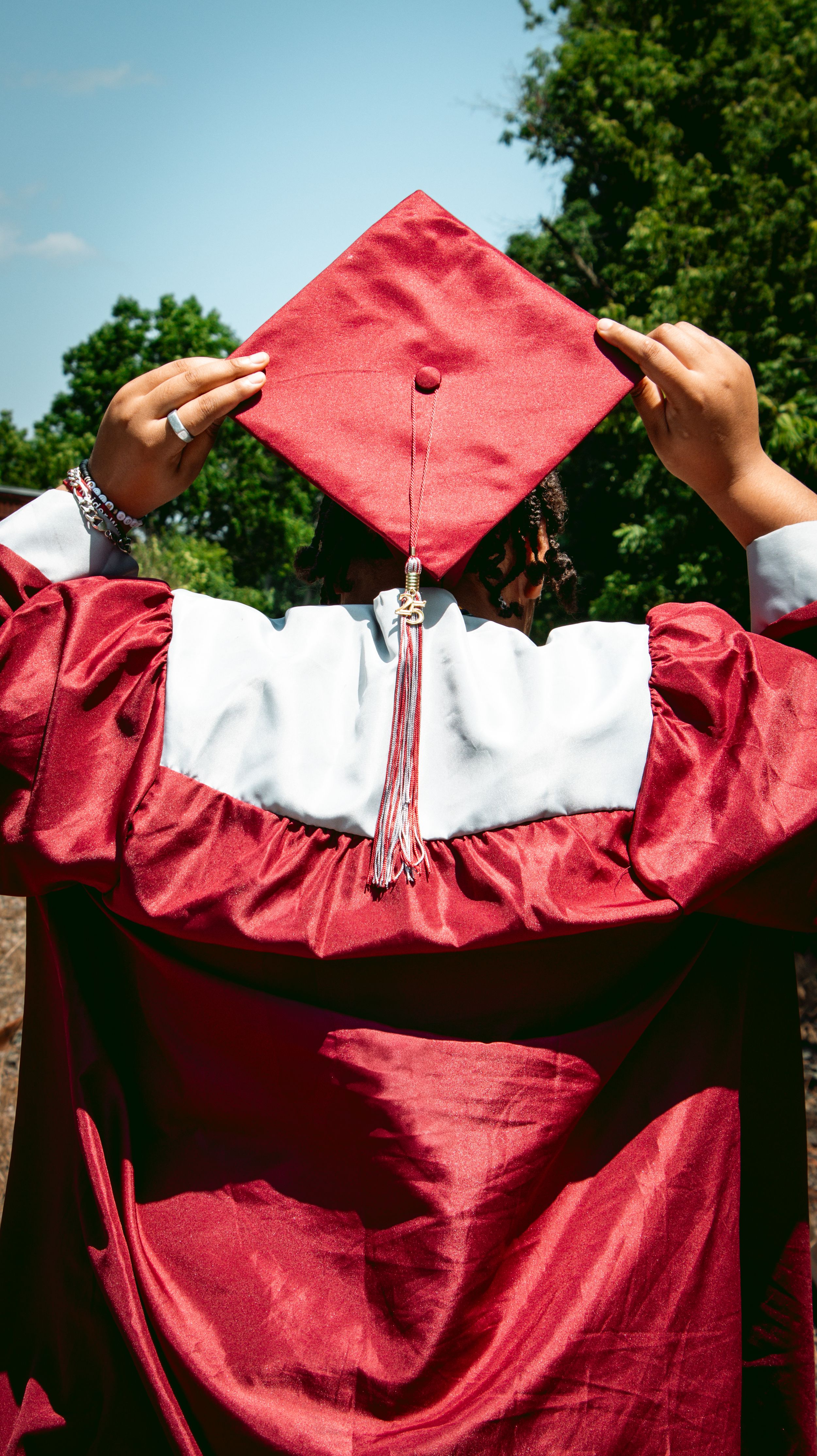 Graduate in red cap and gown holds mortarboard aloft outdoors, viewed from behind with green trees and blue sky.