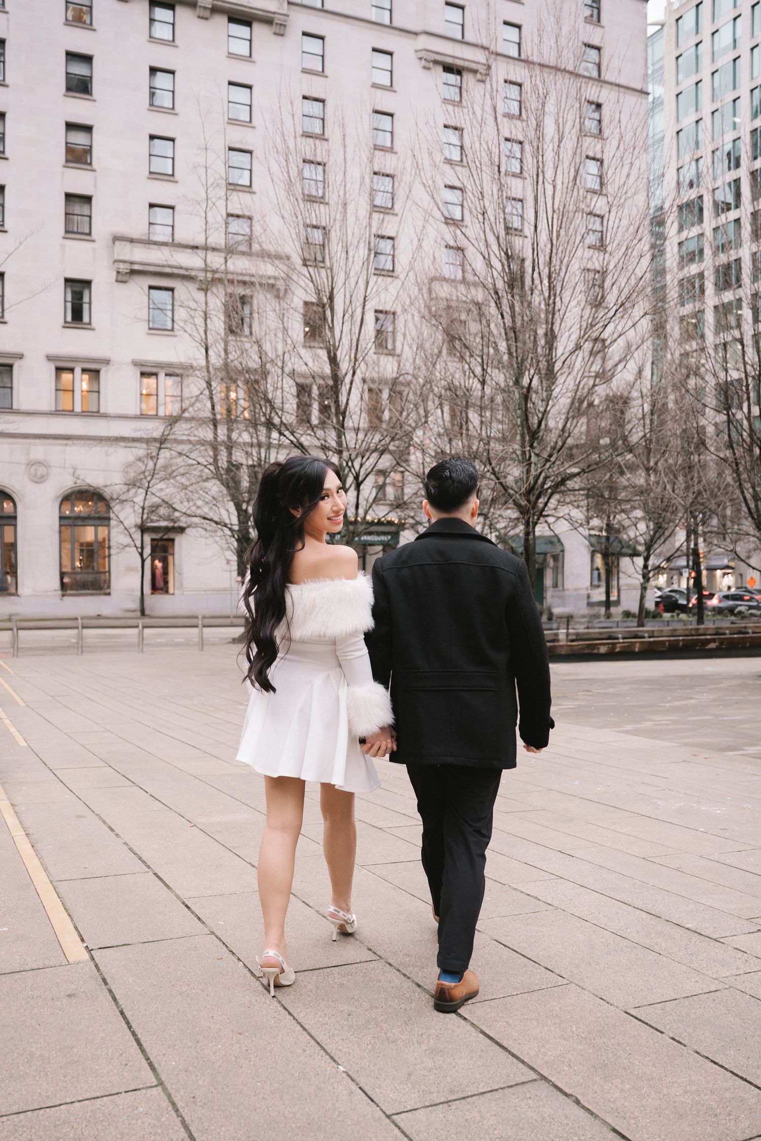 Couple walking hand in hand through an urban plaza, woman in white off-shoulder dress, man in black suit, bare trees behind.