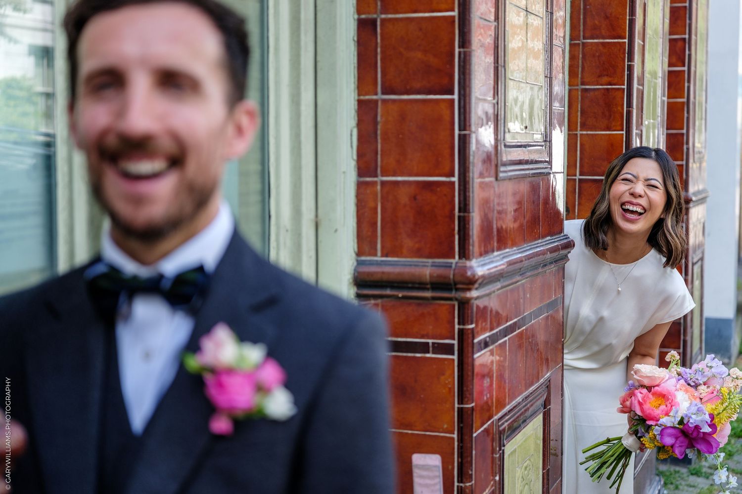 Bride laughing joyfully behind a brick column as groom stands in foreground during a wedding first look moment.
