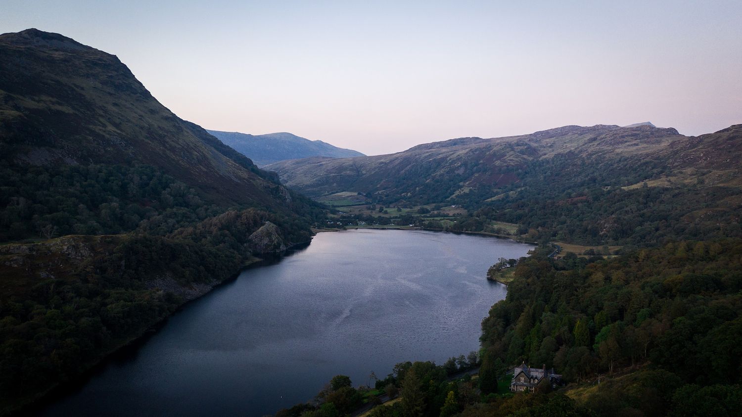 Aerial view of a serene mountain lake surrounded by forested hills and valleys at dusk in a scenic landscape.