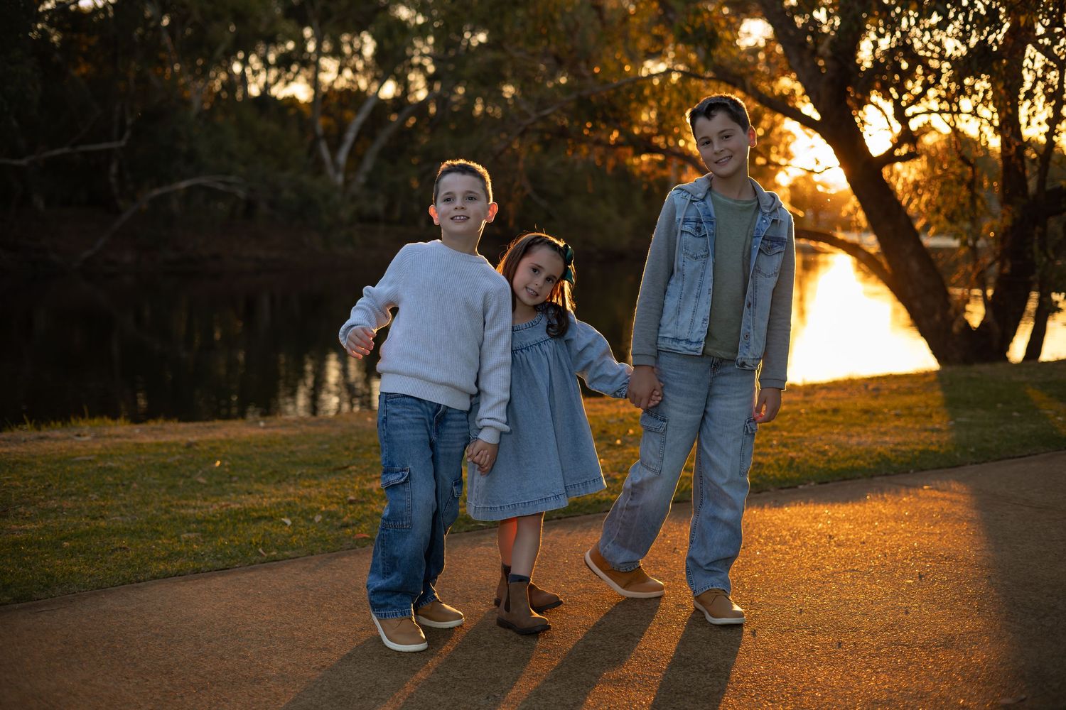Three children holding hands at sunset by a lake in a park, wearing denim and casual outfits.