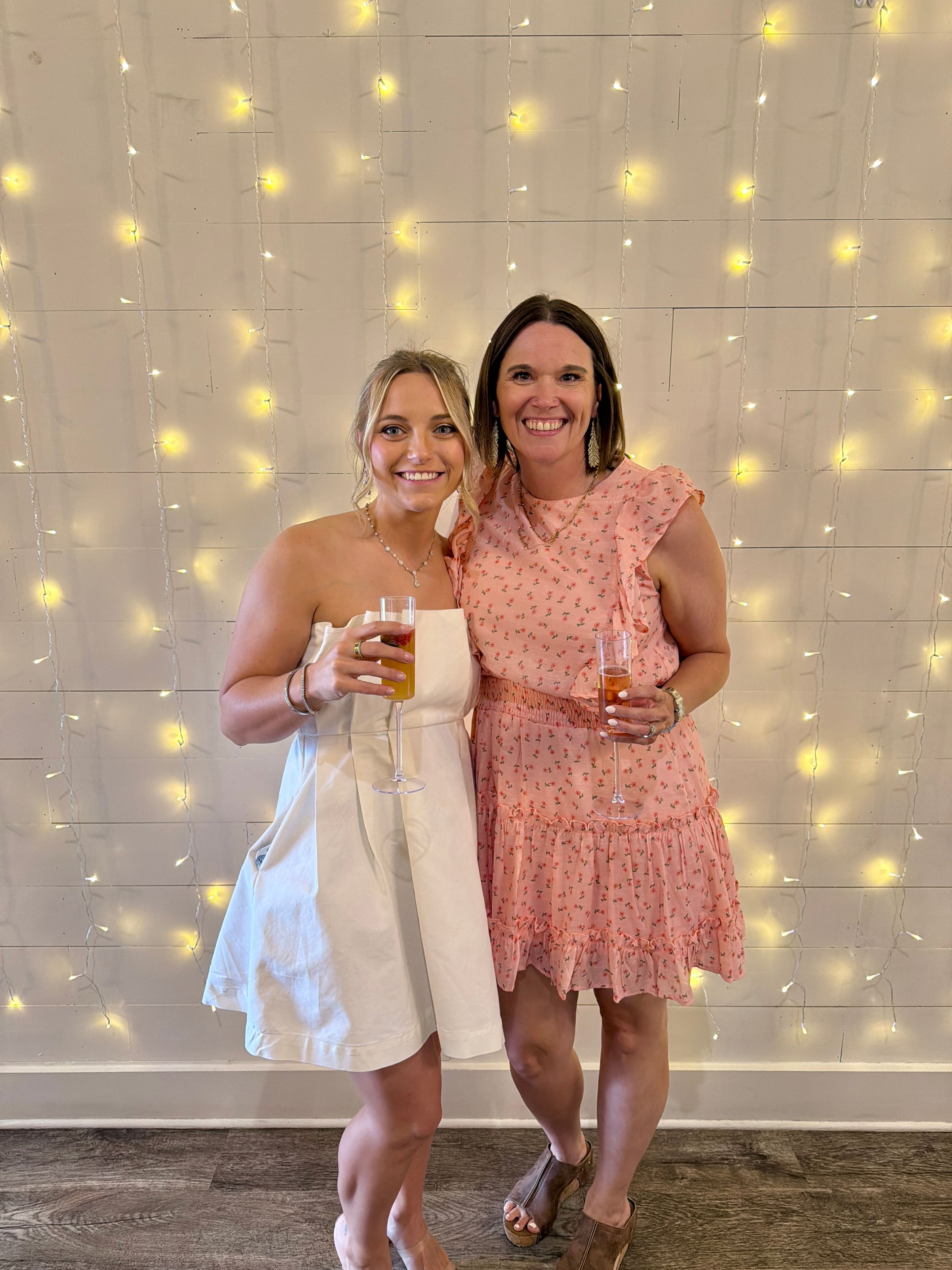 Two smiling women in summer dresses holding drinks pose together in front of a twinkling fairy light backdrop.
