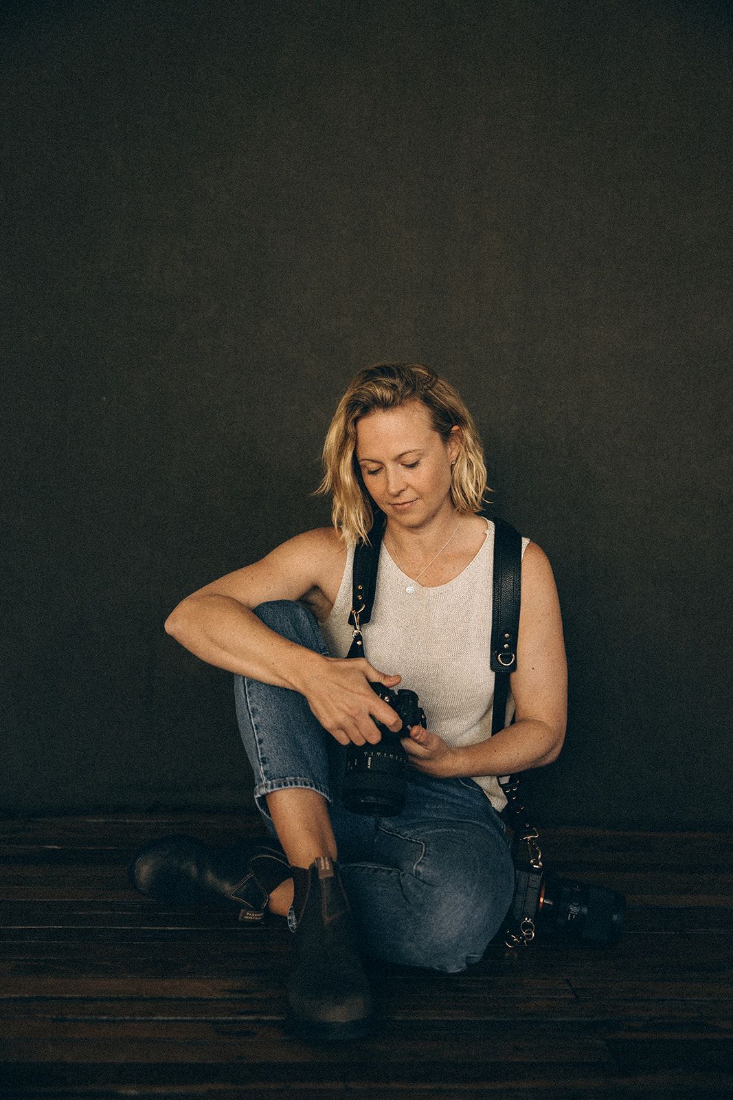 A blonde woman sits on a wooden floor holding a camera, wearing jeans and a white tank top against a dark backdrop.