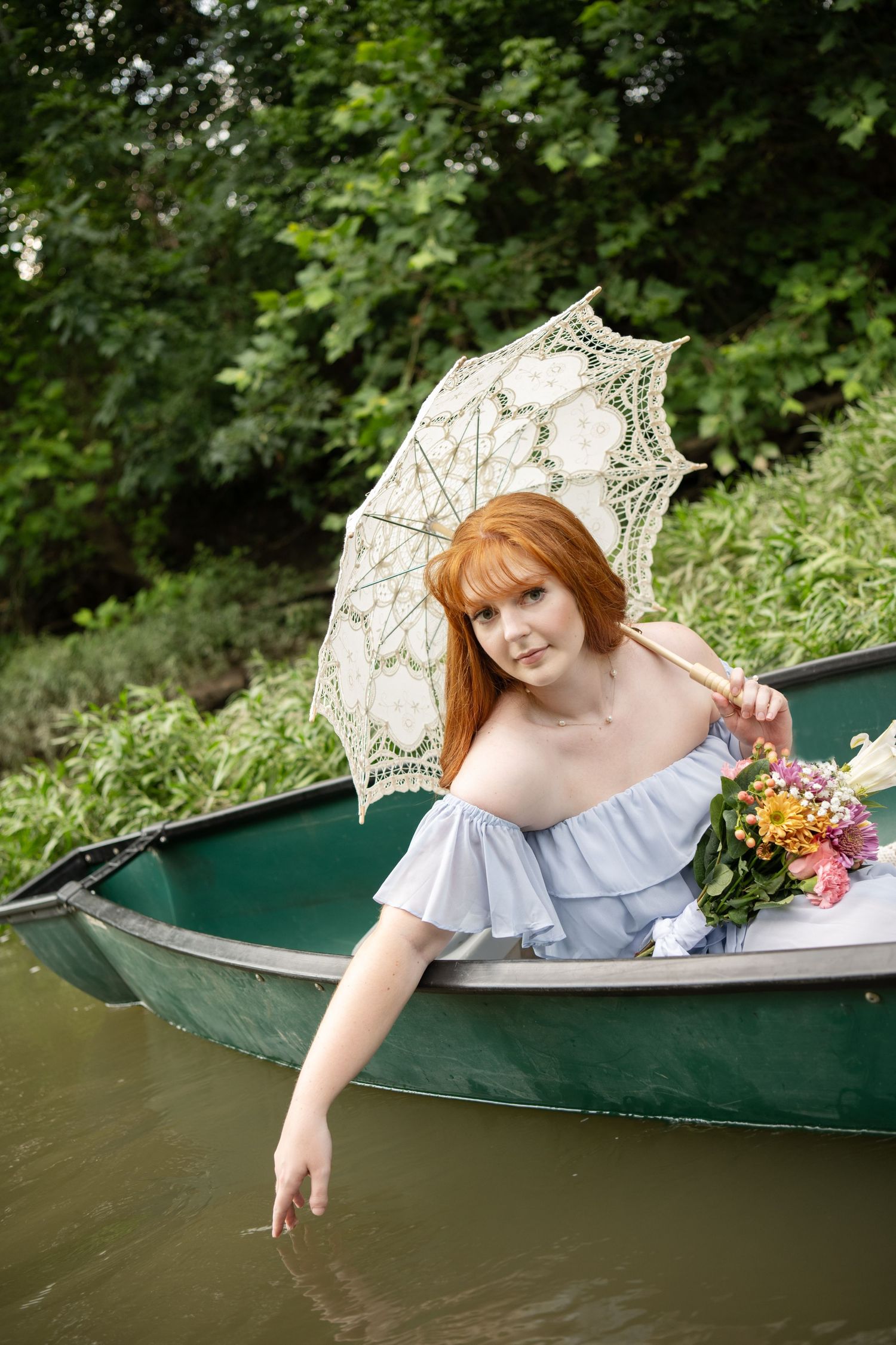 Red-haired woman in blue off-shoulder dress holding lace parasol and flowers while sitting in a green rowboat.