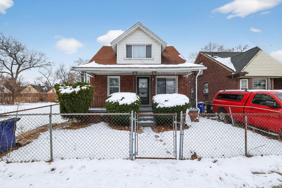 Brick bungalow home with snow-covered yard, chain-link fence, and dormer window under a partly cloudy winter sky.