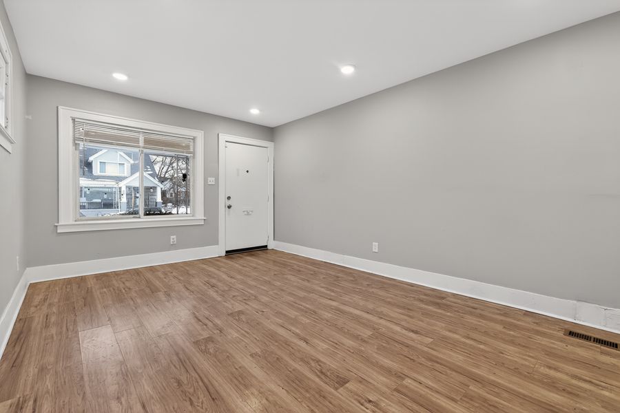 Bright empty living room with light gray walls, hardwood floors, recessed lighting, and a white front door with window.