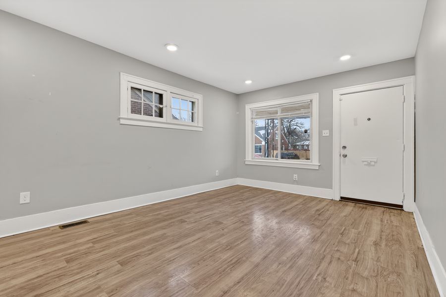 Bright empty living room with light gray walls, hardwood floors, recessed lighting, and a white front door with natural light.