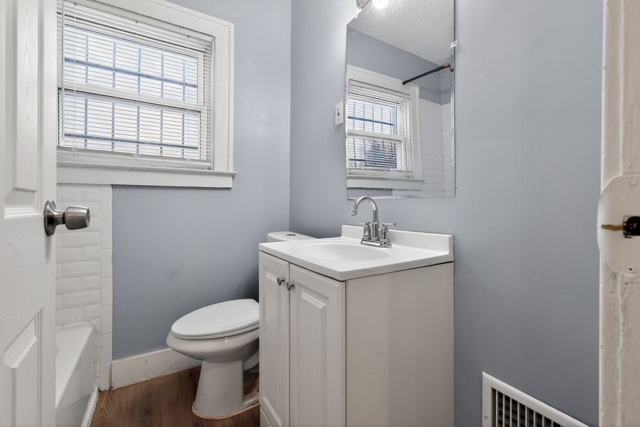 Small bathroom with light blue walls, white vanity, toilet, hardwood floors, and two windows allowing natural light.