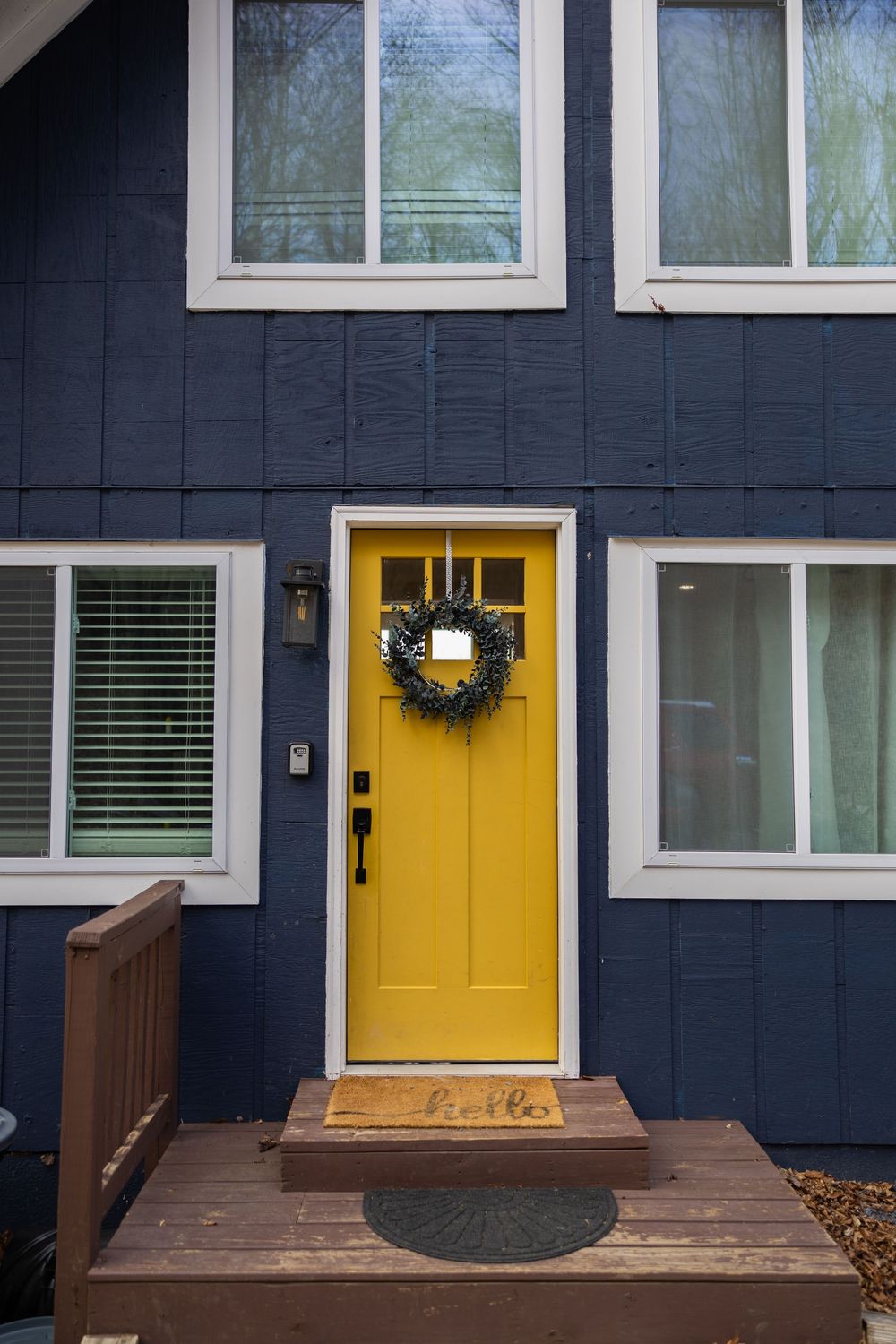 Navy blue home exterior with bold yellow front door adorned with wreath, white trim windows, and a hello doormat on wood porch.