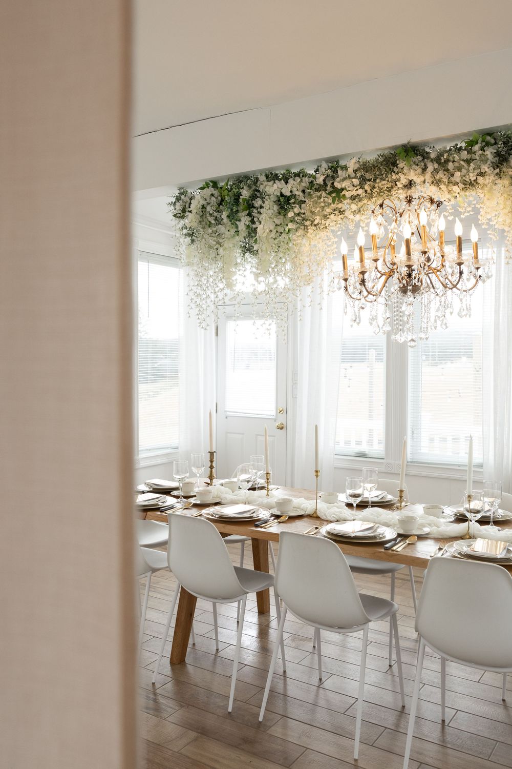 Elegant dining room with white chairs, wooden table, gold candelabras, crystal chandelier, and cascading white floral ceiling installation.