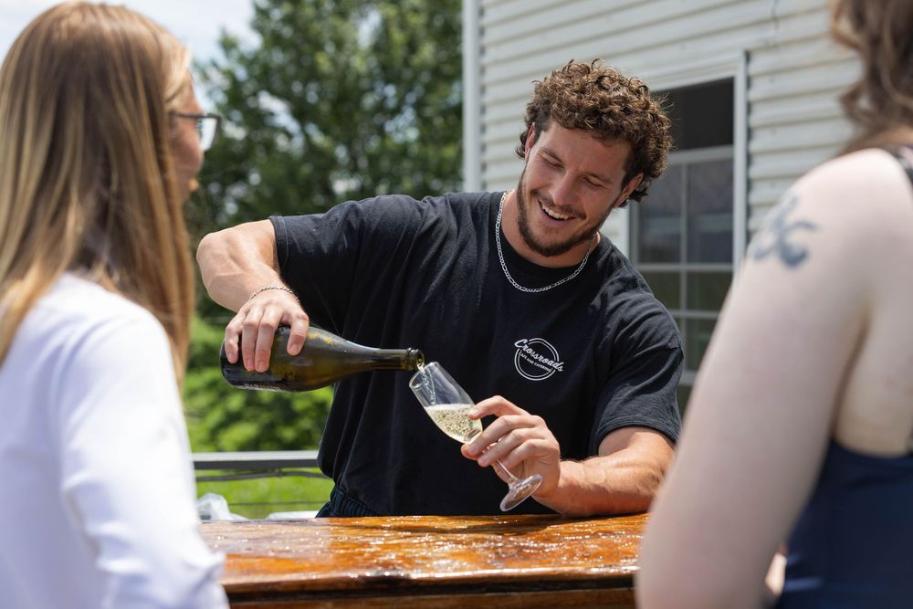 Man smiling while pouring champagne into a glass outdoors at a sunny backyard gathering with two women.