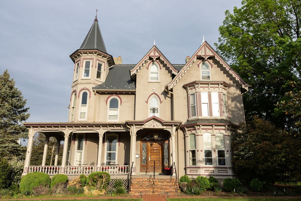 Victorian mansion with turret, wraparound porch, ornate trim, and bay windows surrounded by lush trees at dusk.