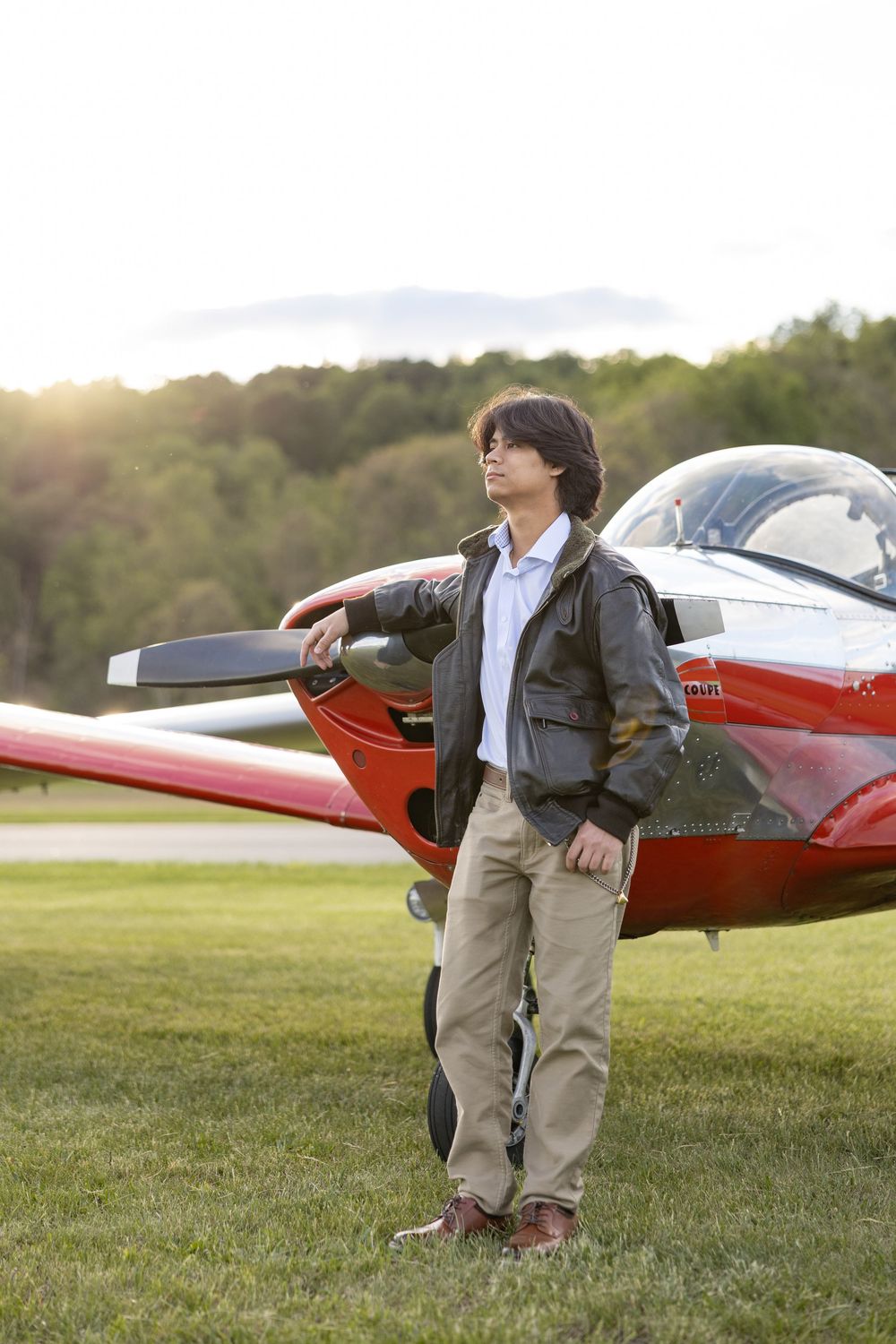 Young pilot in flight jacket leaning on red propeller plane on grassy airfield at sunset.