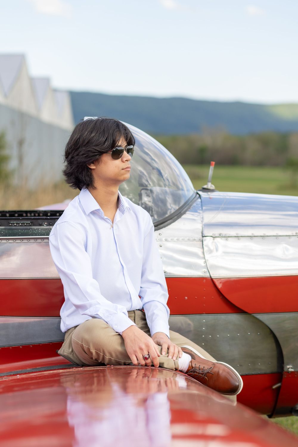 Young man in sunglasses and white shirt leaning on a red and silver small aircraft outdoors.