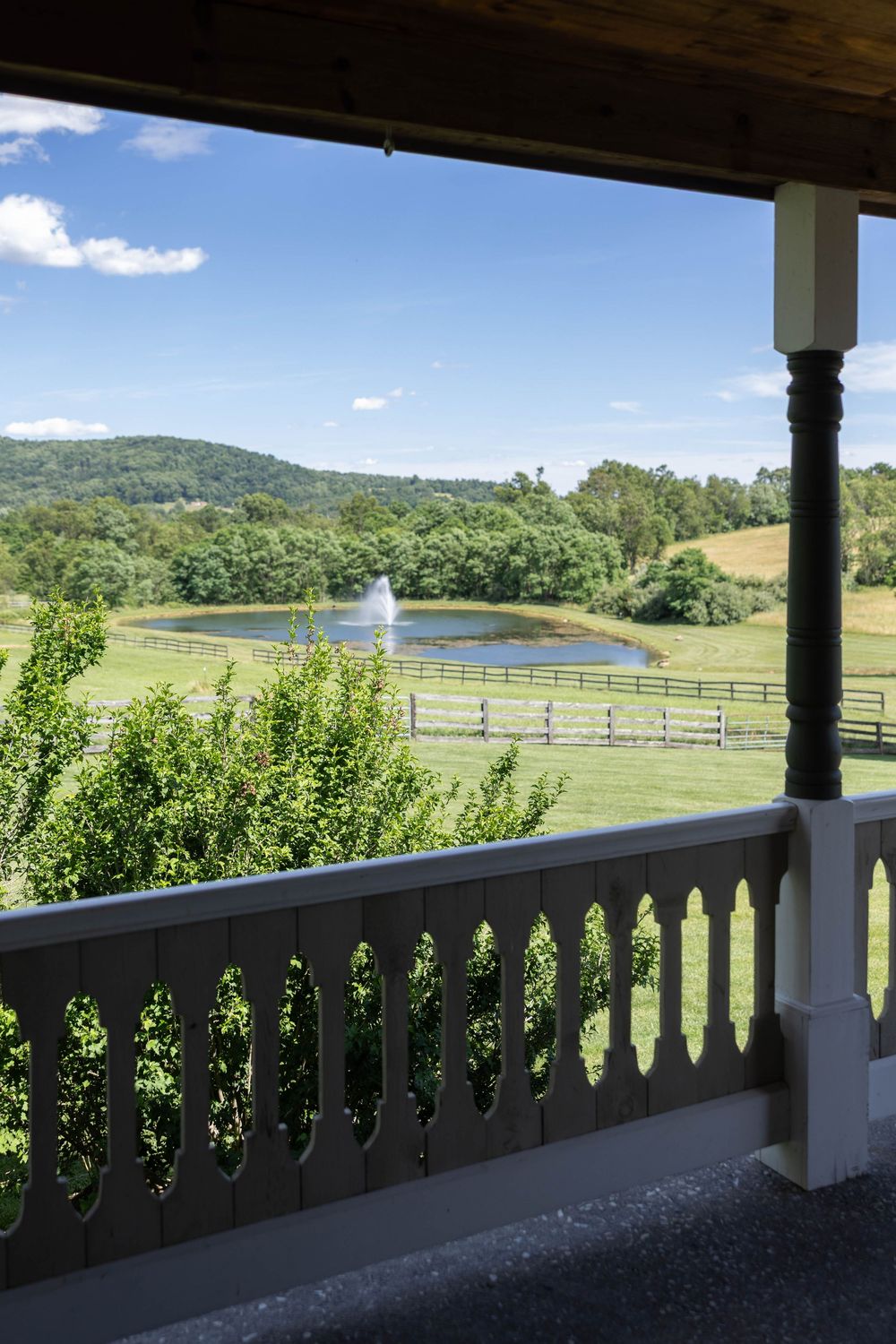 Scenic porch view overlooking a pond with fountain, green pastures, wooden fences, and rolling hills under a blue sky.