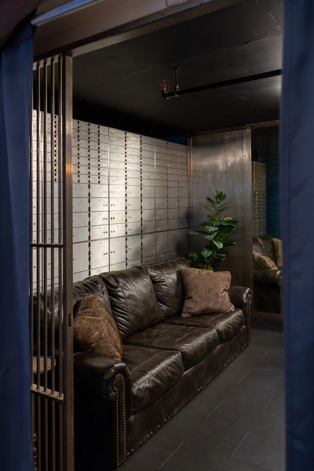 Luxury bank vault lounge with leather sofa, decorative pillows, safety deposit boxes, and a fiddle leaf fig plant.