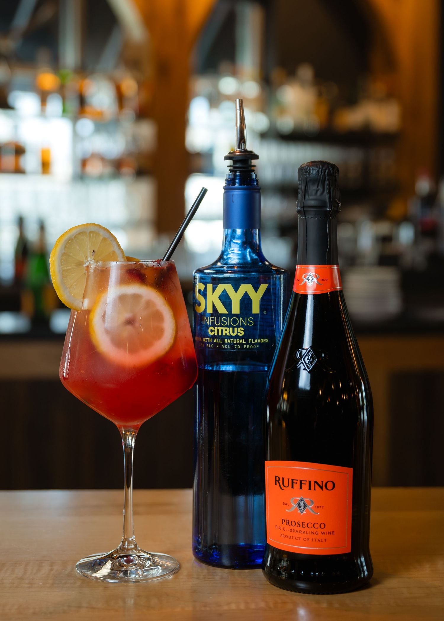 A citrus cocktail garnished with lemon beside SKYY Infusions Citrus vodka and Ruffino Prosecco bottles on a bar.