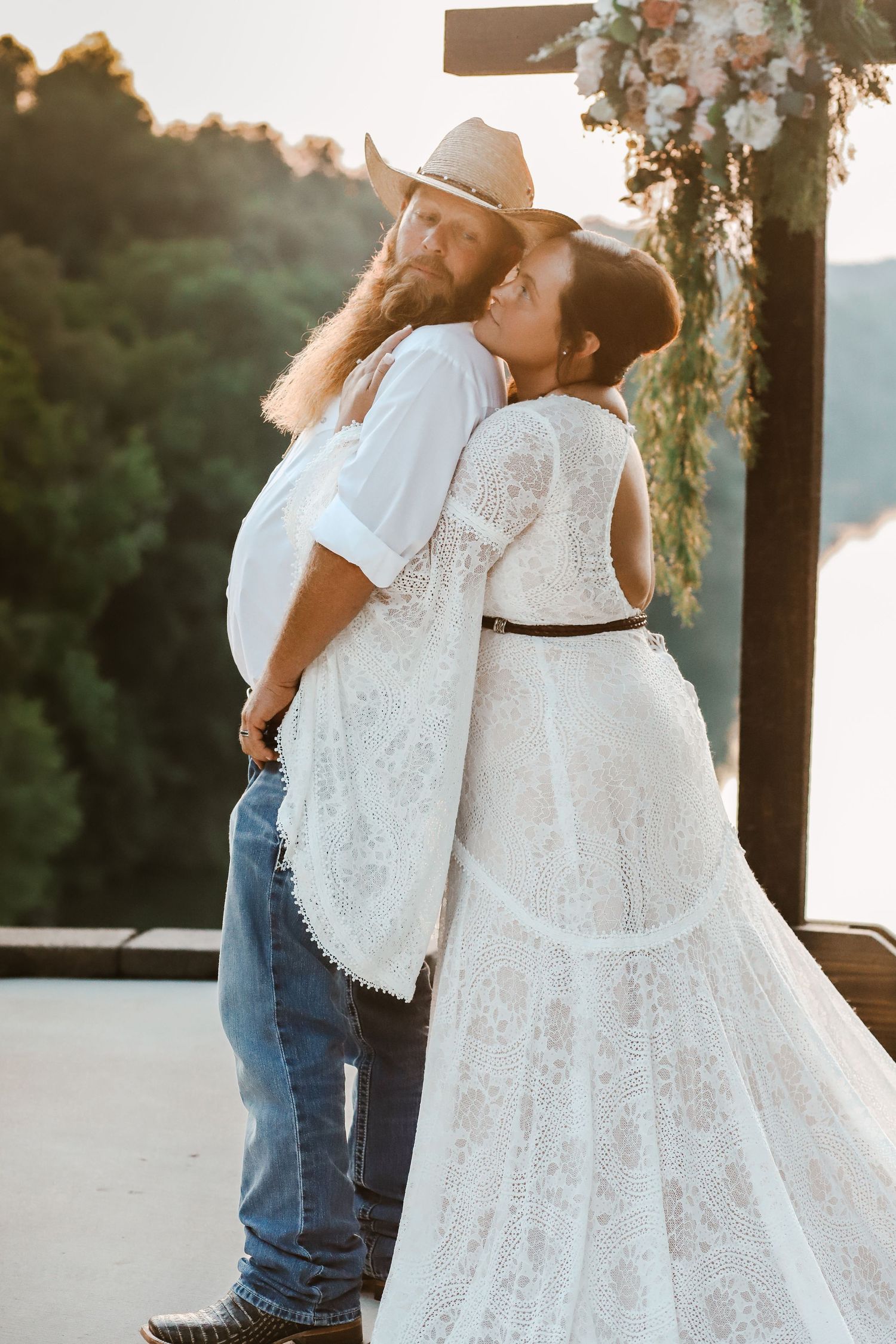 Bride in lace boho wedding dress embraces bearded groom in cowboy hat and jeans at outdoor lakeside ceremony.
