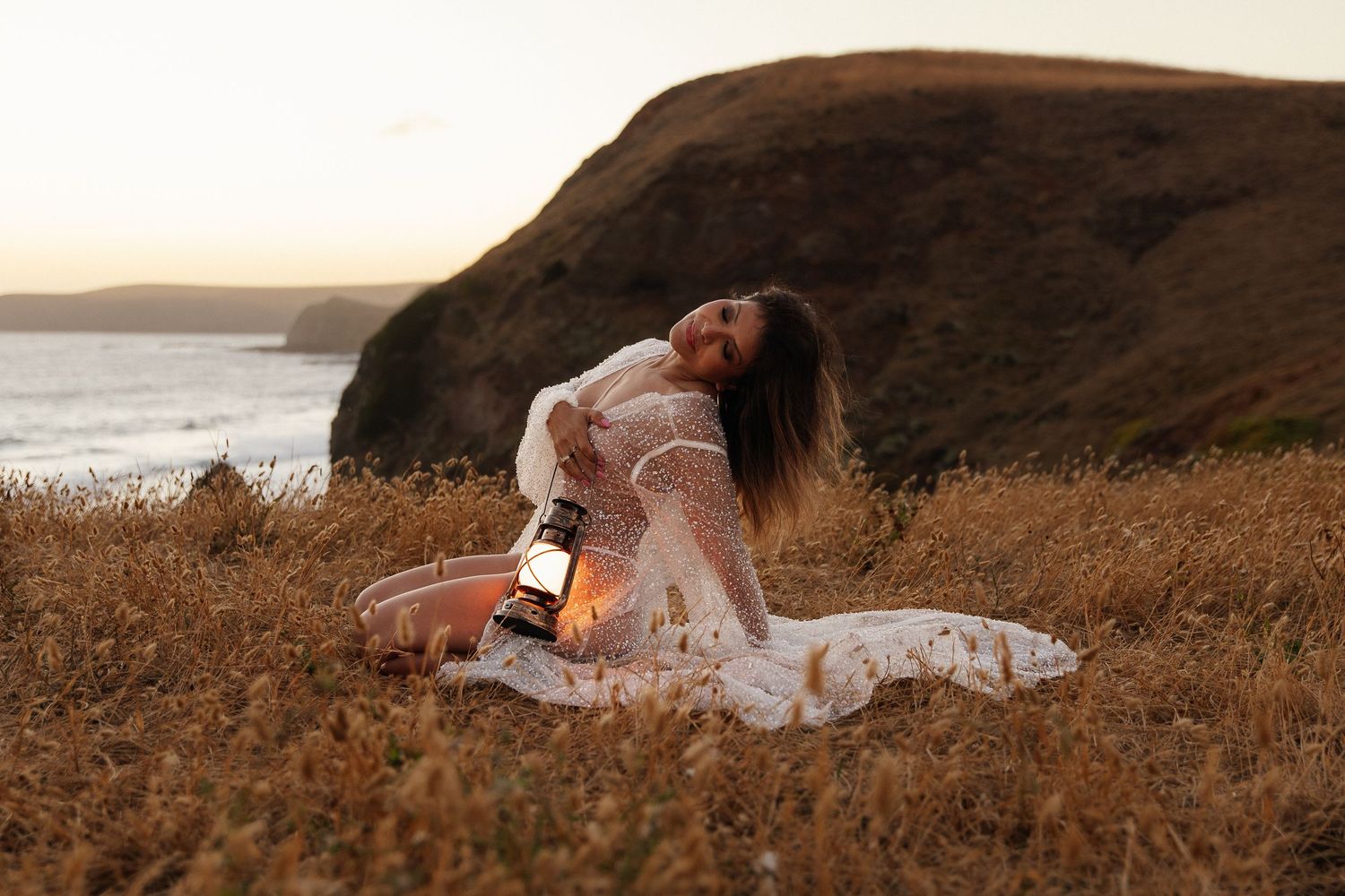 Woman in white lace dress sitting in golden grass at sunset, holding a lantern near coastal cliffs.