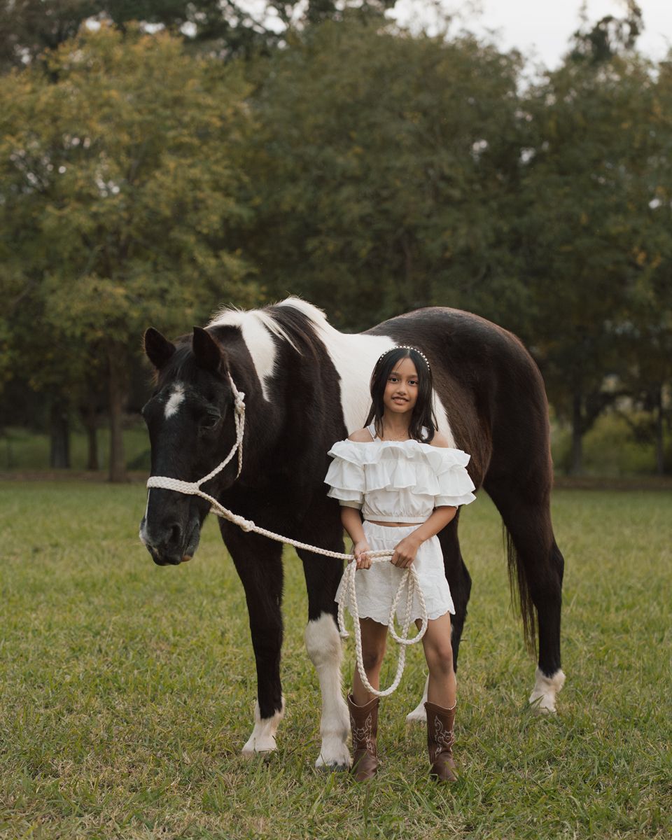 A girl in white dress and brown boots holds rope lead of black and white paint horse in green field in Sydney family photoshoot with a horse