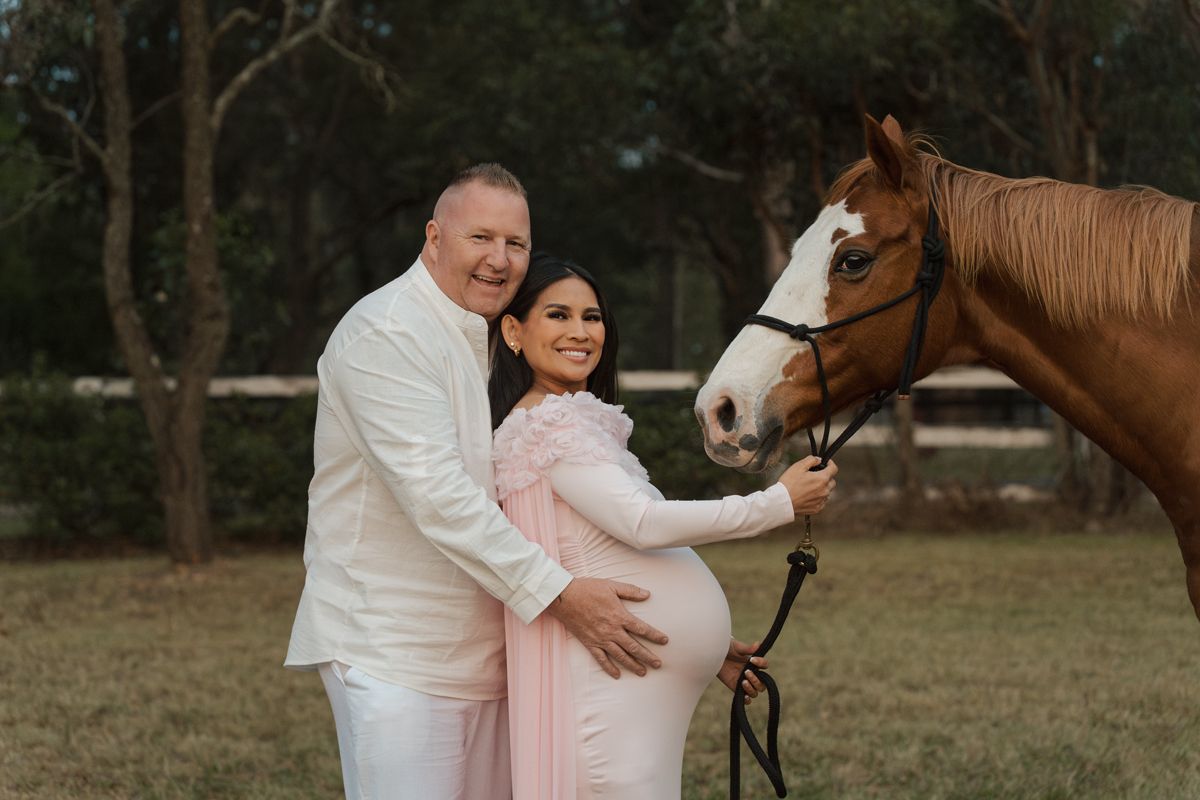 Couple in white and pink maternity outfits posing with a brown horse outdoors during a maternity photoshoot with a horse in Sydney