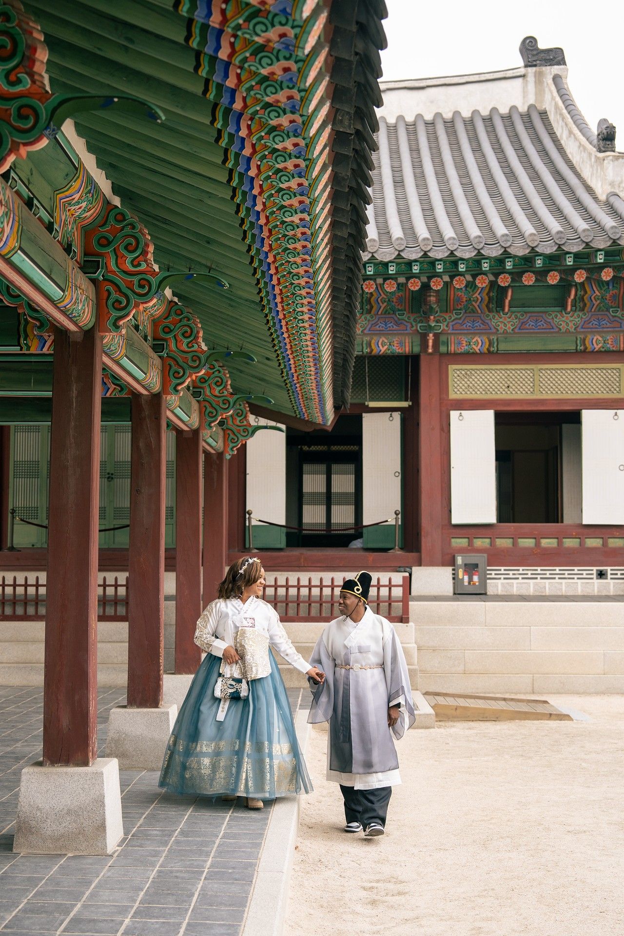 Couple in traditional Korean hanbok attire walking hand in hand at Gyeongbokgung Palace courtyard.