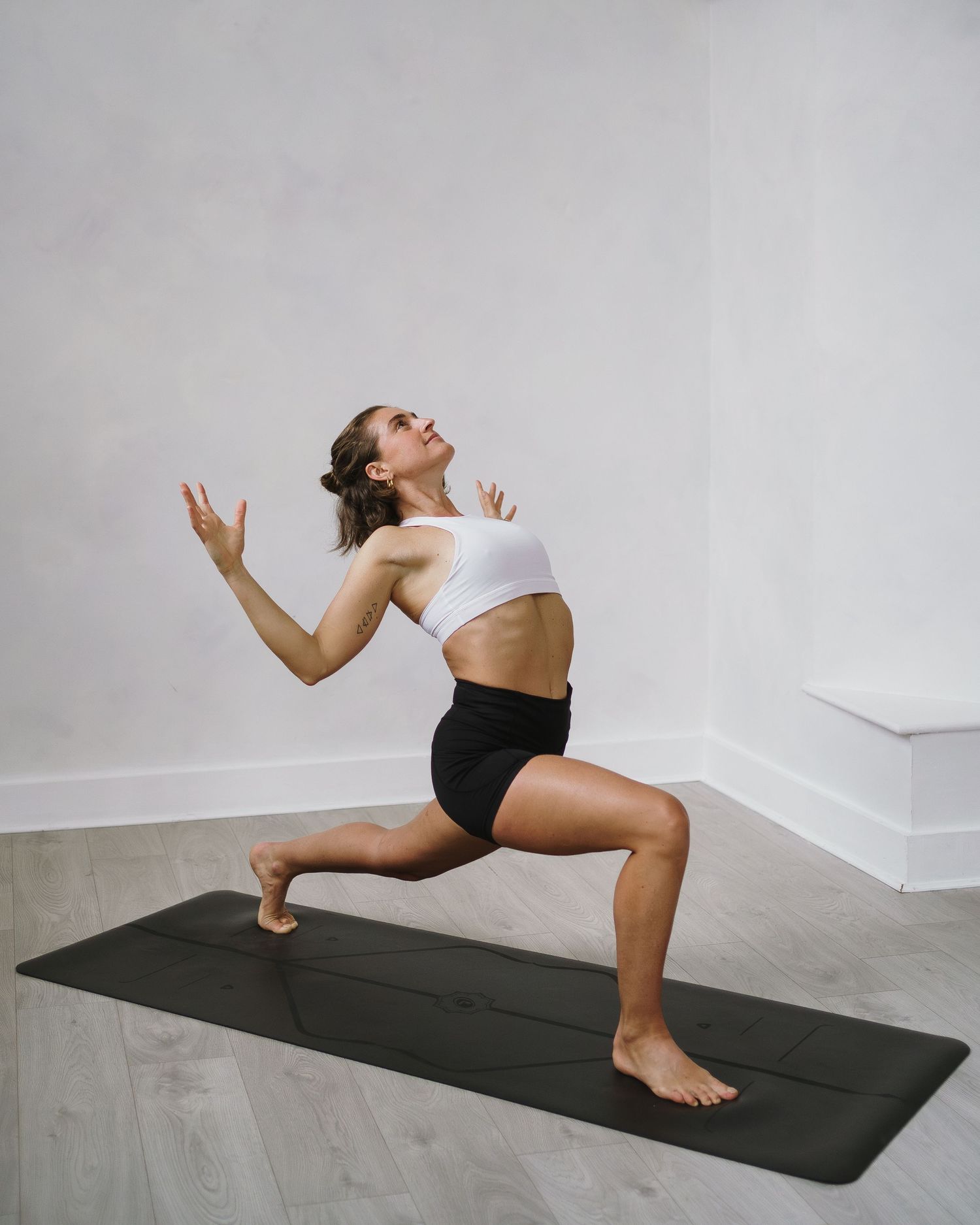 Woman performing a lunge pose on a yoga mat wearing white crop top and black shorts in a minimalist studio setting.