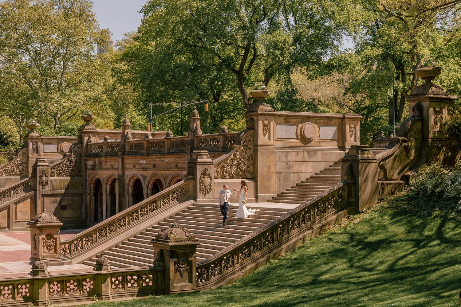 Couple in wedding attire on ornate stone staircase at Bethesda Terrace in Central Park, NYC, surrounded by lush green trees.