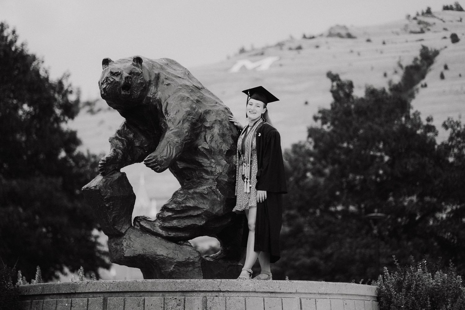 Graduate in cap and gown poses beside a large bear statue outdoors in a black and white photo.