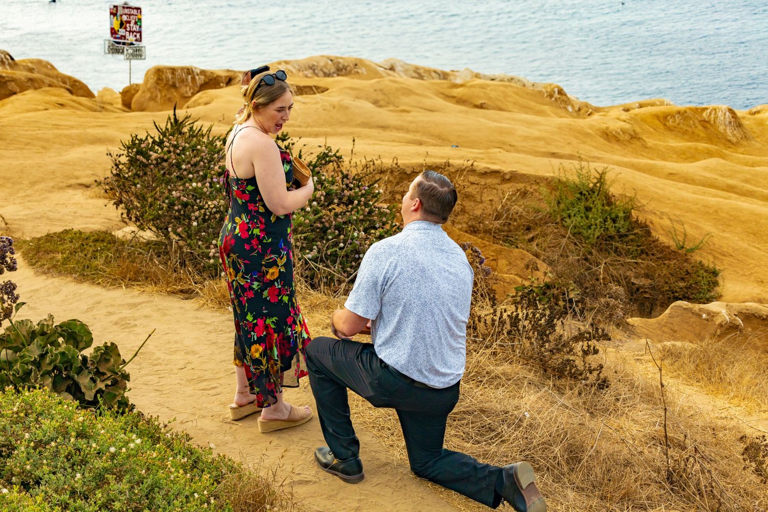 Man proposes on one knee to woman on scenic coastal cliffside trail with golden rocks and ocean view.