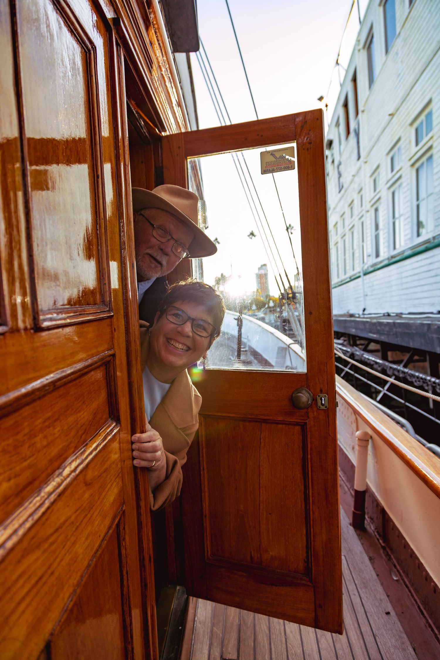 Two smiling people peek through a wooden door on a vintage boat deck, with sunlight streaming behind them.