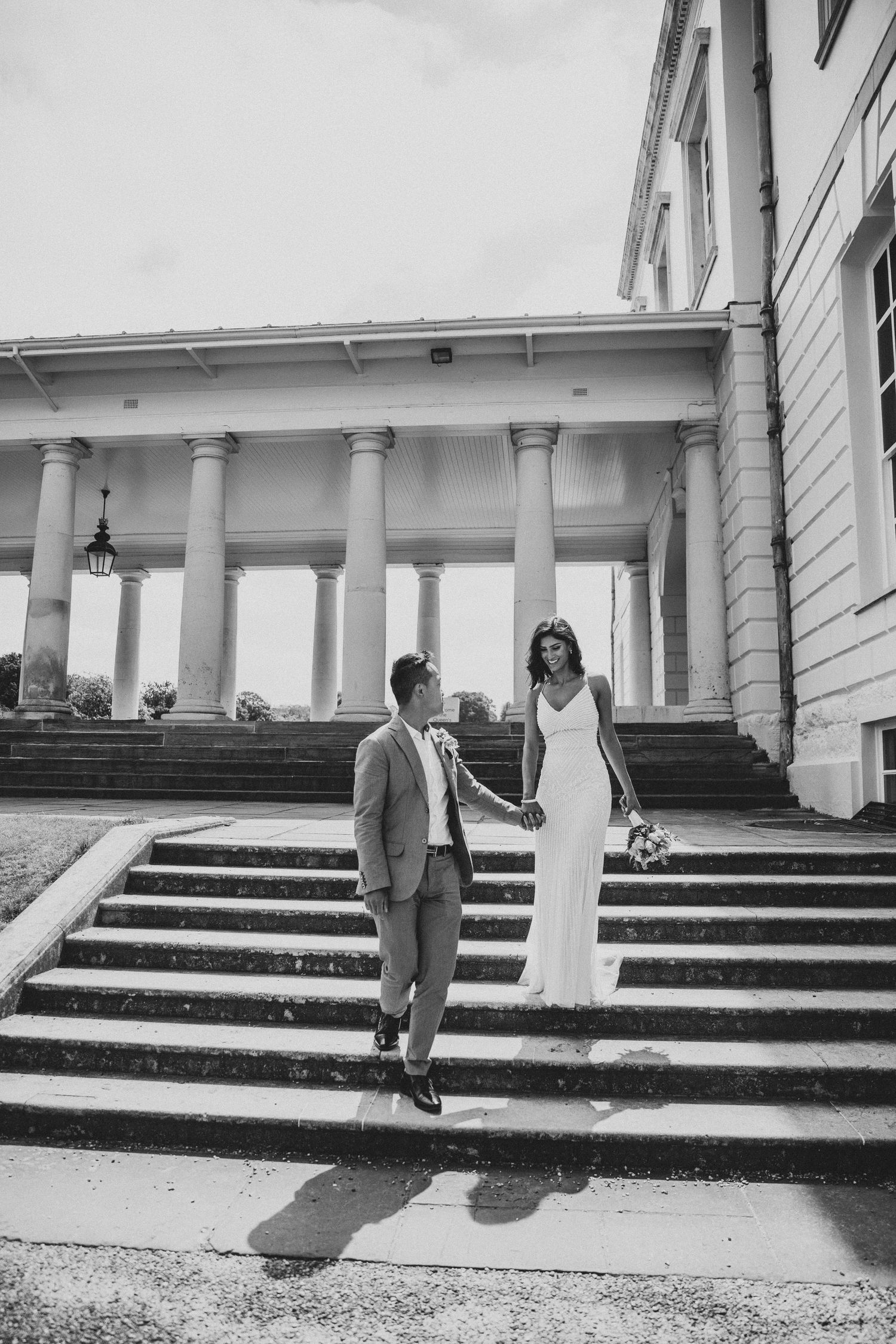 Black and white photo of a bride and groom holding hands on steps outside a grand columned building.