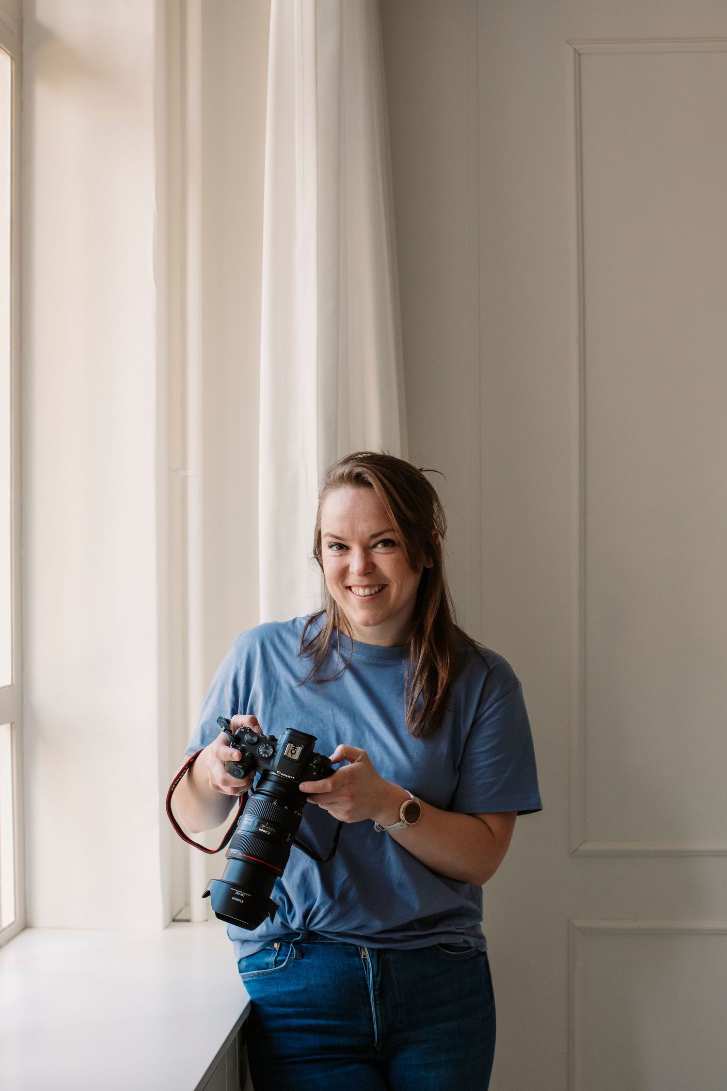 Photographer in blue t-shirt holds professional camera by window in bright room.
