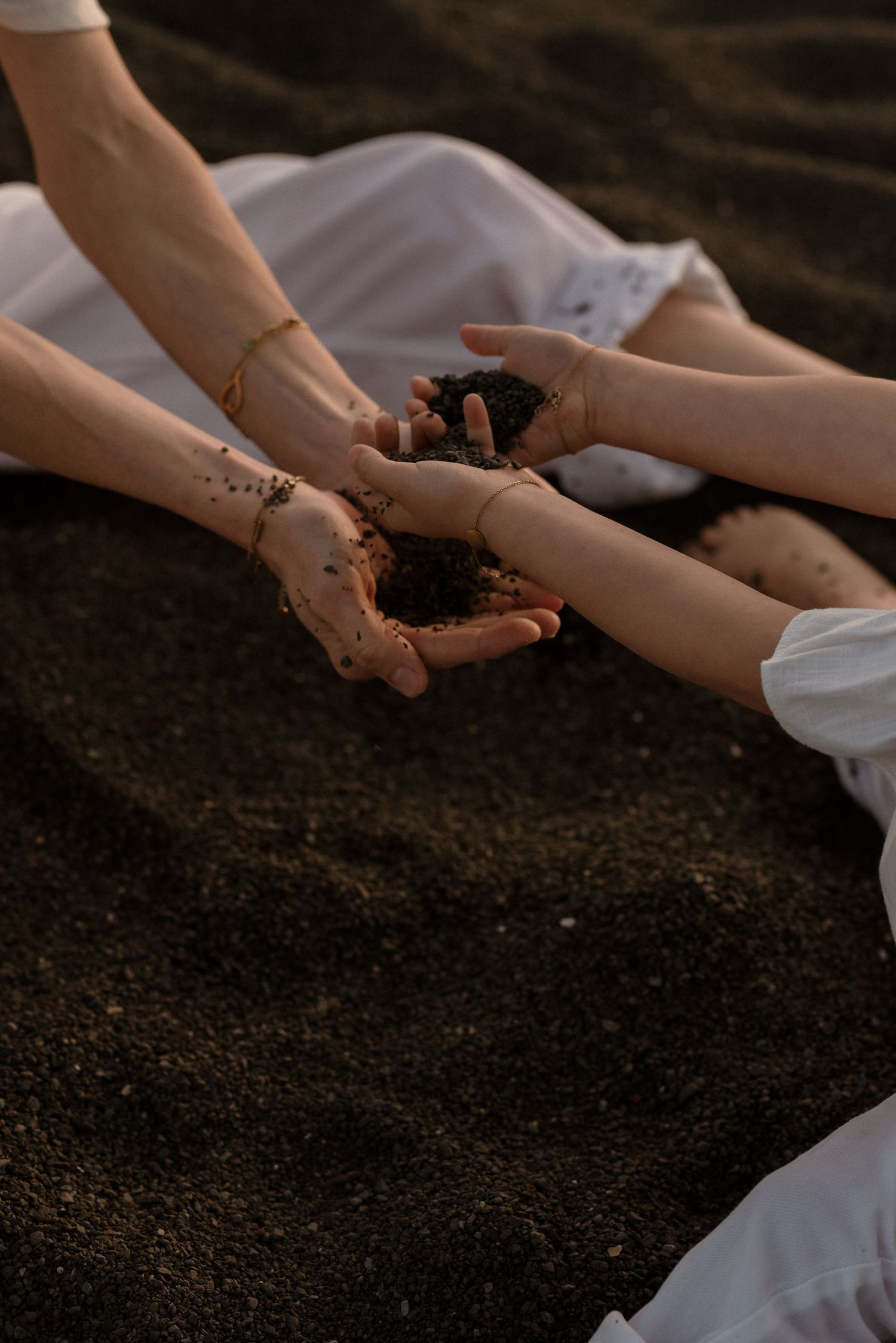 Two people in white dresses holding dark sand in their hands on a black sand beach.